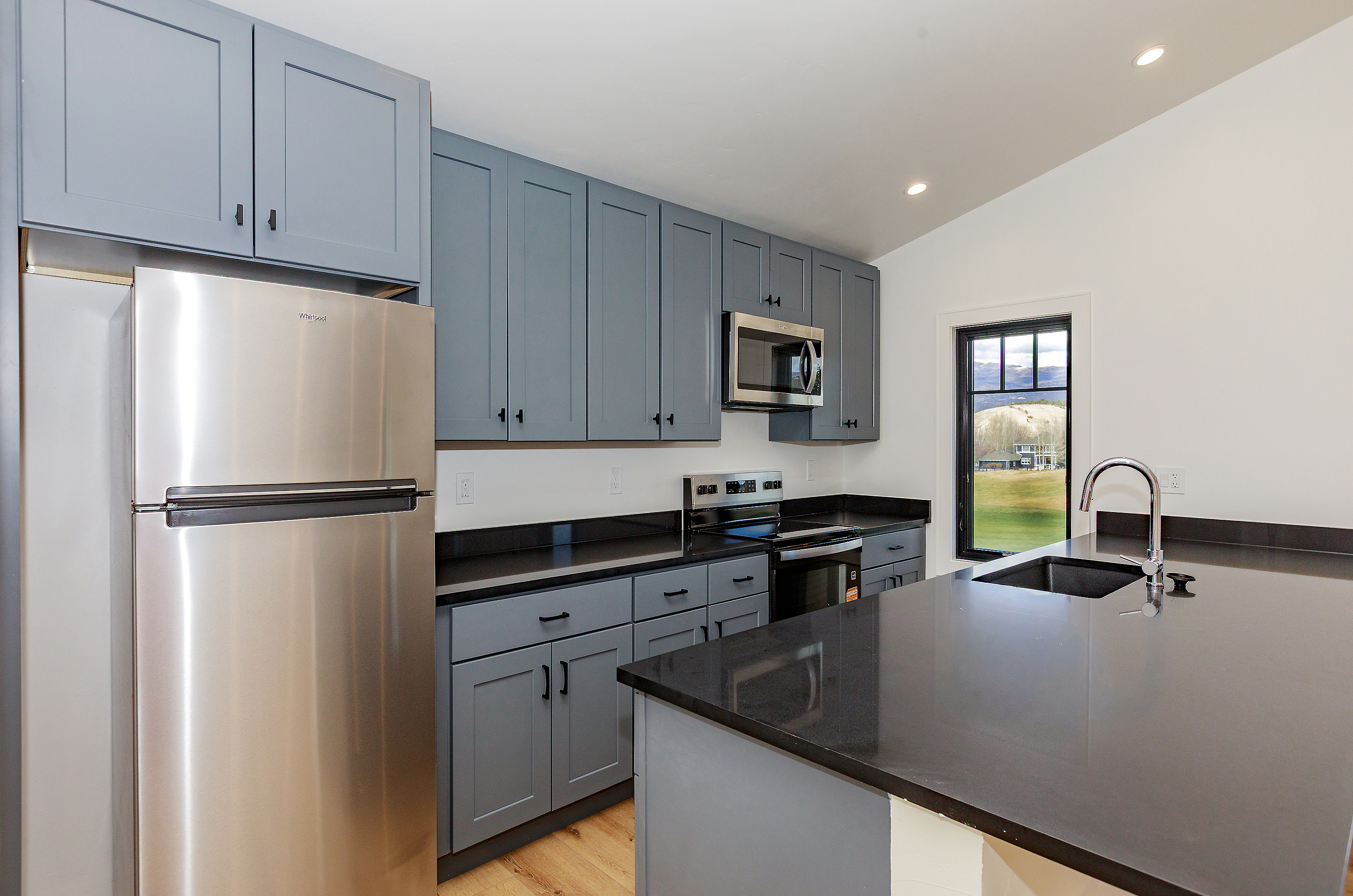 This is a well-lit kitchen featuring gray cabinets with black hardware and black countertops. A stainless steel refrigerator is on the left, and a stainless steel microwave and oven are visible along the back wall. An island with a sink and faucet is in the foreground, and a window provides a view of the outside.