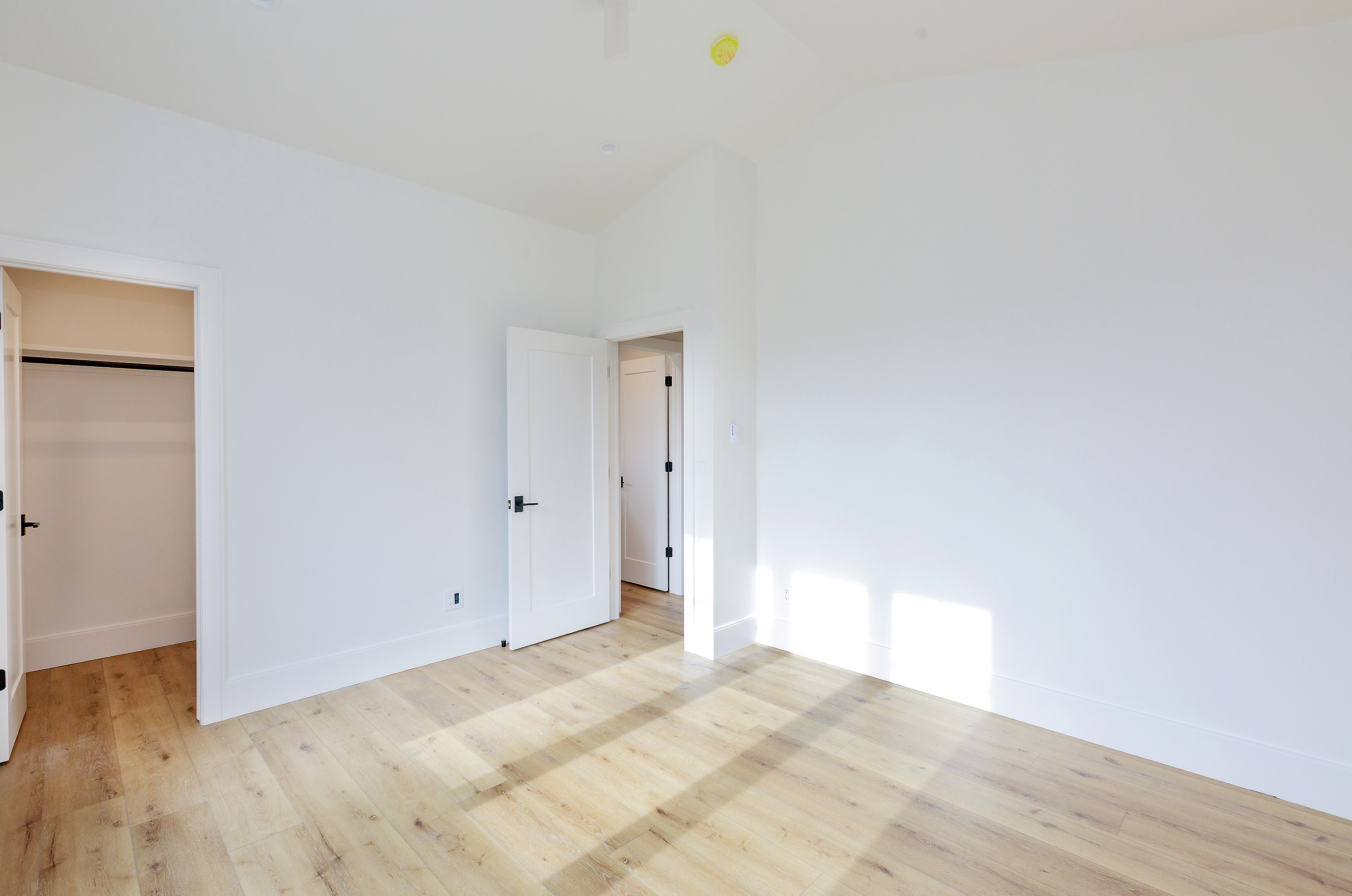 This is a bright and airy bedroom featuring light wood flooring and white walls, creating a clean and modern aesthetic. The room has multiple doorways, one leading to a closet and another to an adjacent space, enhancing the sense of openness. Natural light floods the room, highlighting the minimalist design and spacious feel.
