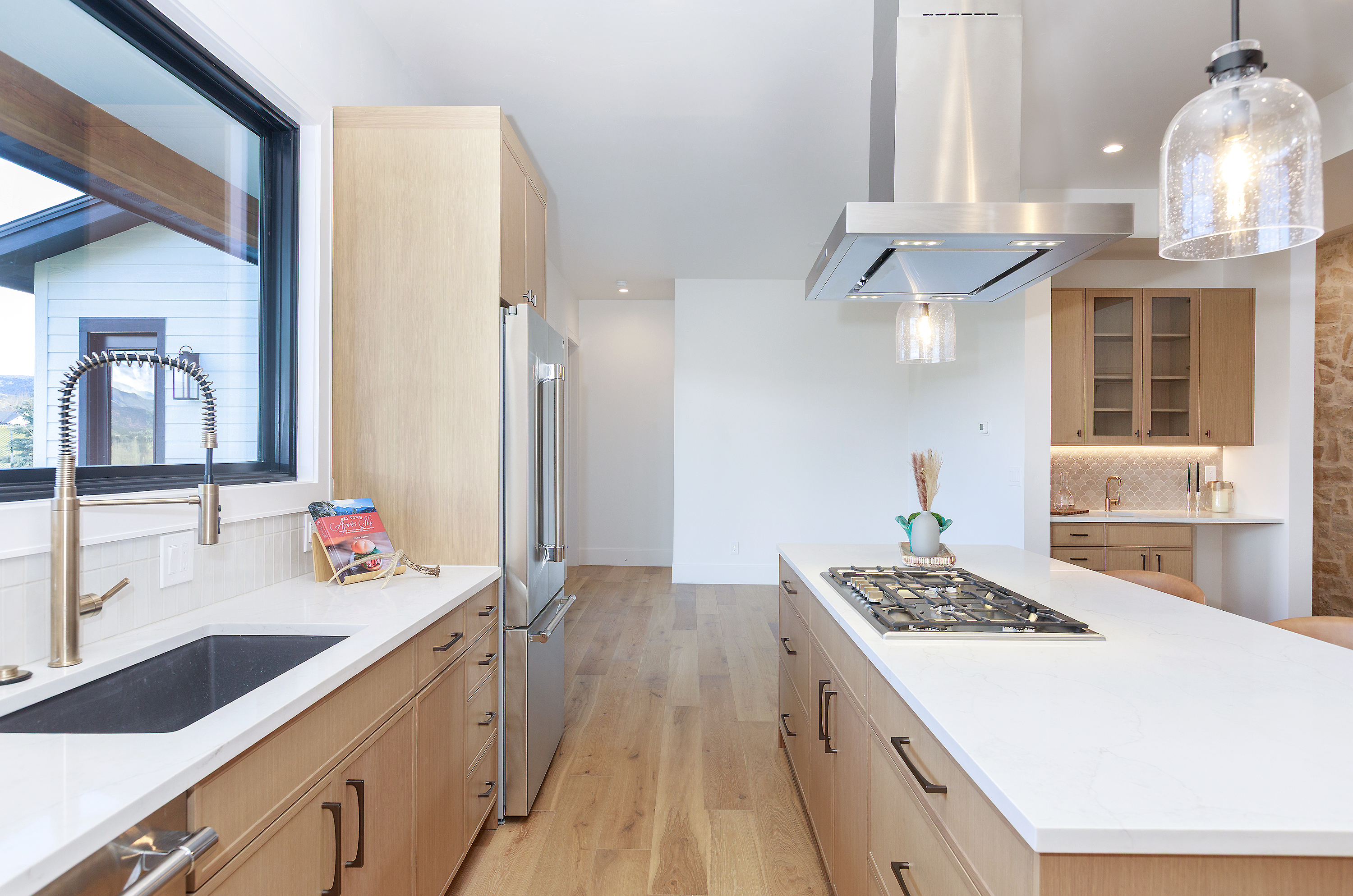 This is a bright and modern kitchen featuring light wood cabinetry, stainless steel appliances, and a large island with a white countertop and built-in gas cooktop. A stainless steel range hood hangs above the island, and a large window provides natural light and a view of the exterior. The kitchen has a clean and contemporary style with hardwood flooring.
