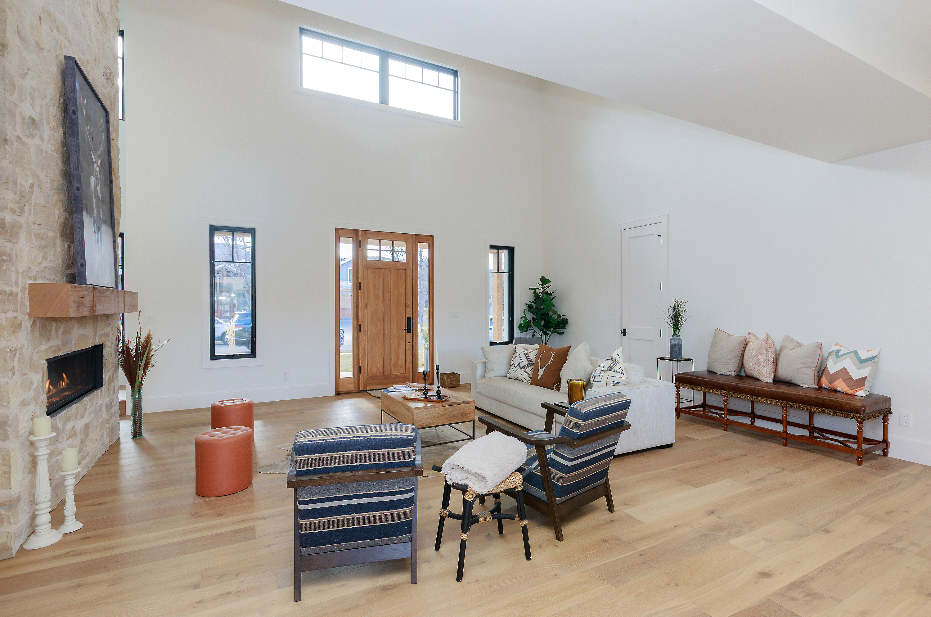 This is an interior shot of a living room featuring a stone fireplace, light wood flooring, and high ceilings. The room is furnished with a light-colored sofa, striped armchairs, and a wooden coffee table. Natural light floods the space through large windows, creating a bright and airy atmosphere.