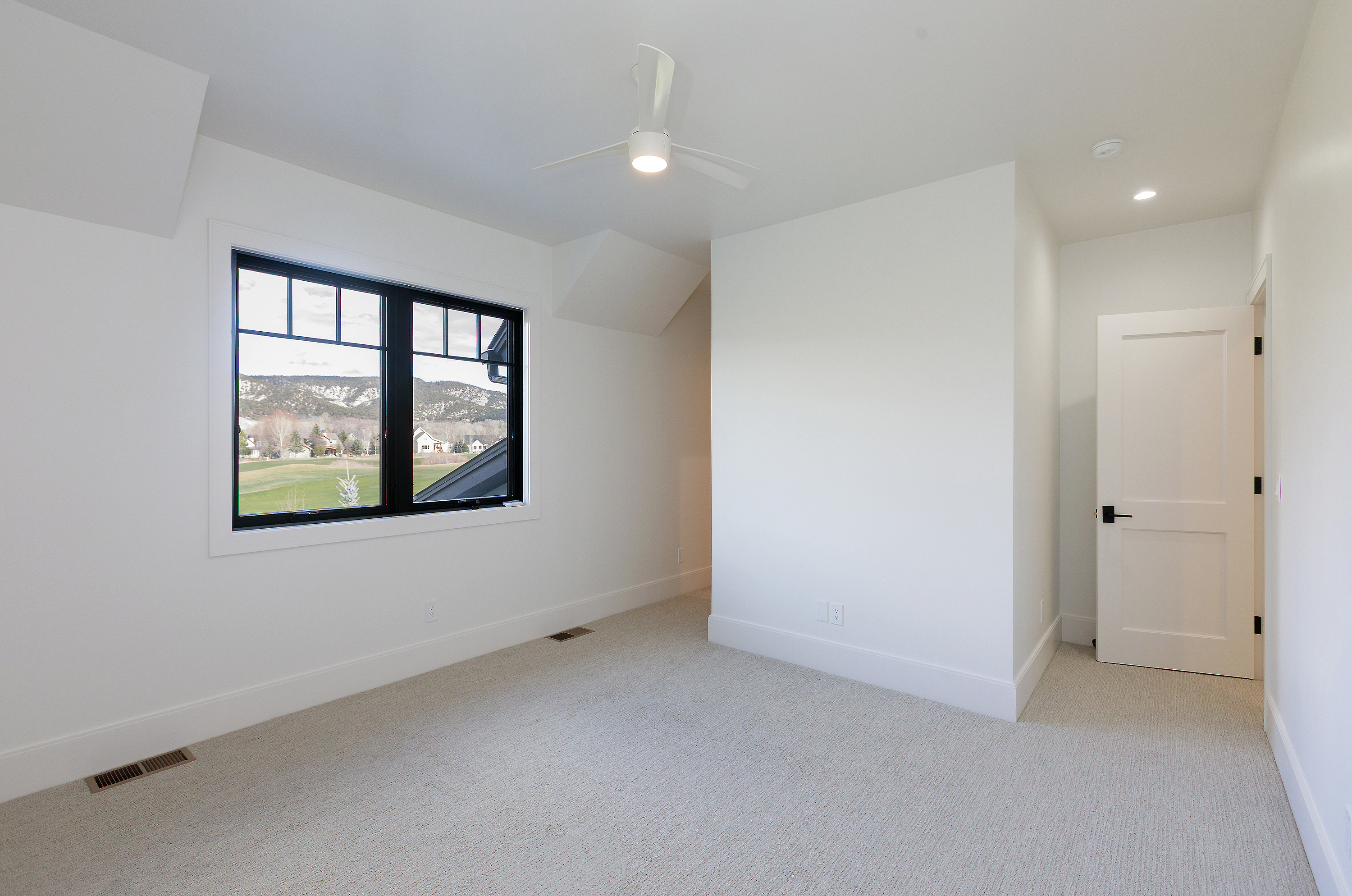 This is a bright, modern bedroom with white walls and light carpet. A large window with a black frame offers a view of the outdoors. The room is minimalist and clean, creating a serene and inviting atmosphere.