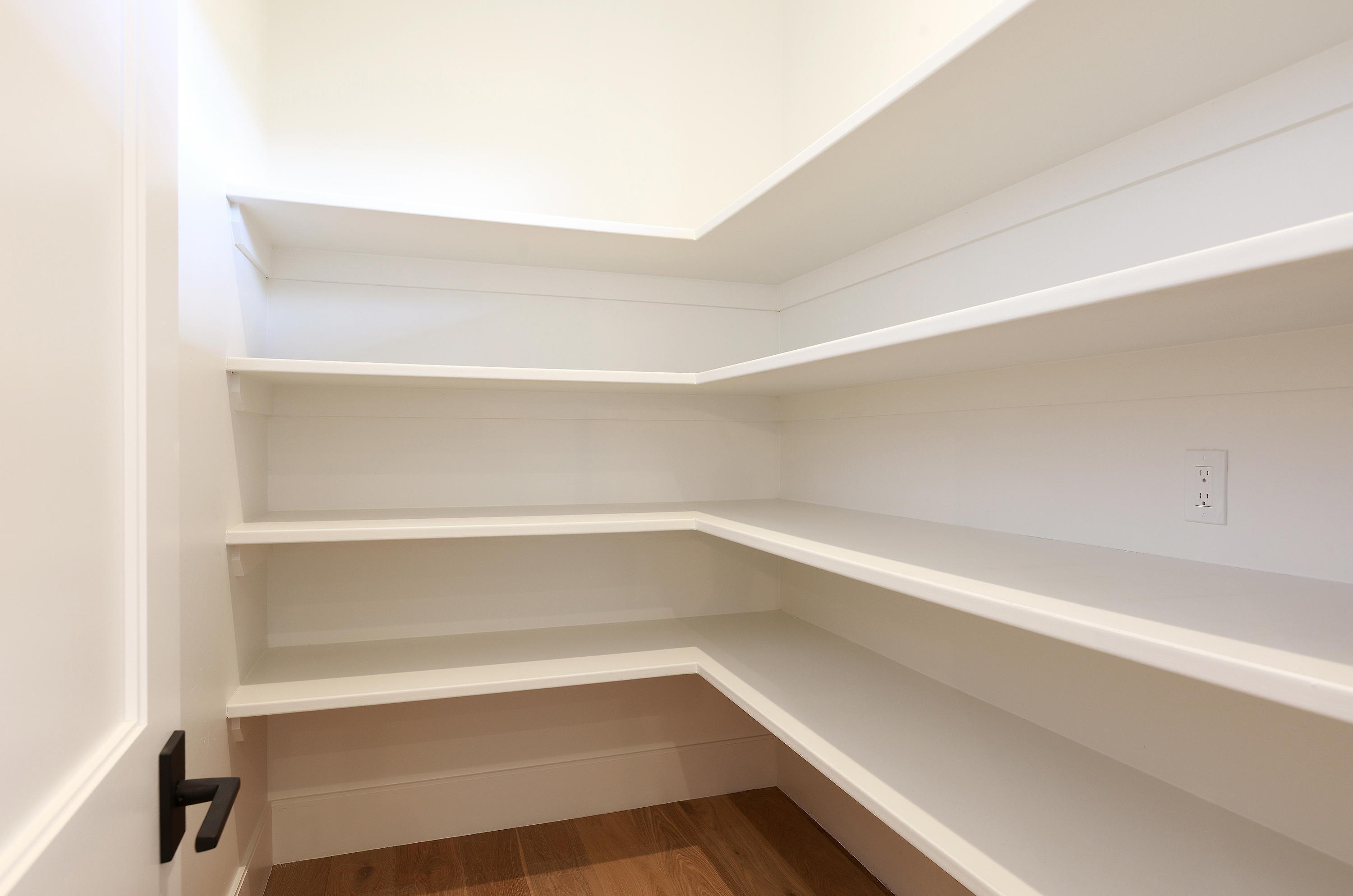 This is an interior shot of a well-organized pantry featuring white shelving. The shelves are spacious and appear to be made of painted wood, offering ample storage space. An electrical outlet is visible on the wall, and the flooring appears to be hardwood, adding a touch of warmth to the otherwise bright and clean space.