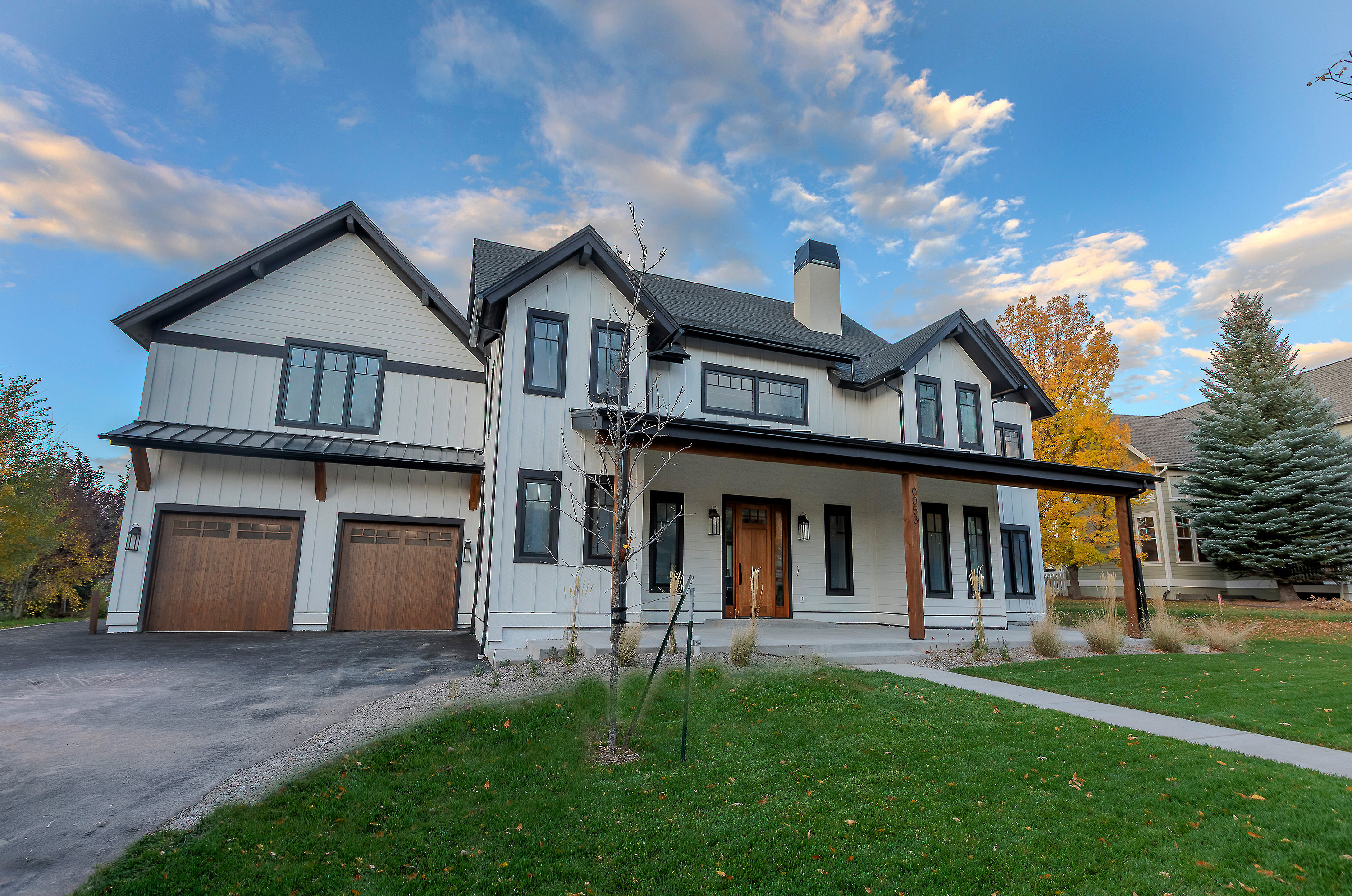 This is a front view of a modern farmhouse-style home. The exterior features white siding with black trim, a covered front porch with wooden support beams, and a well-manicured lawn. The house has a two-car garage with wooden doors and a combination of vertical and horizontal siding.