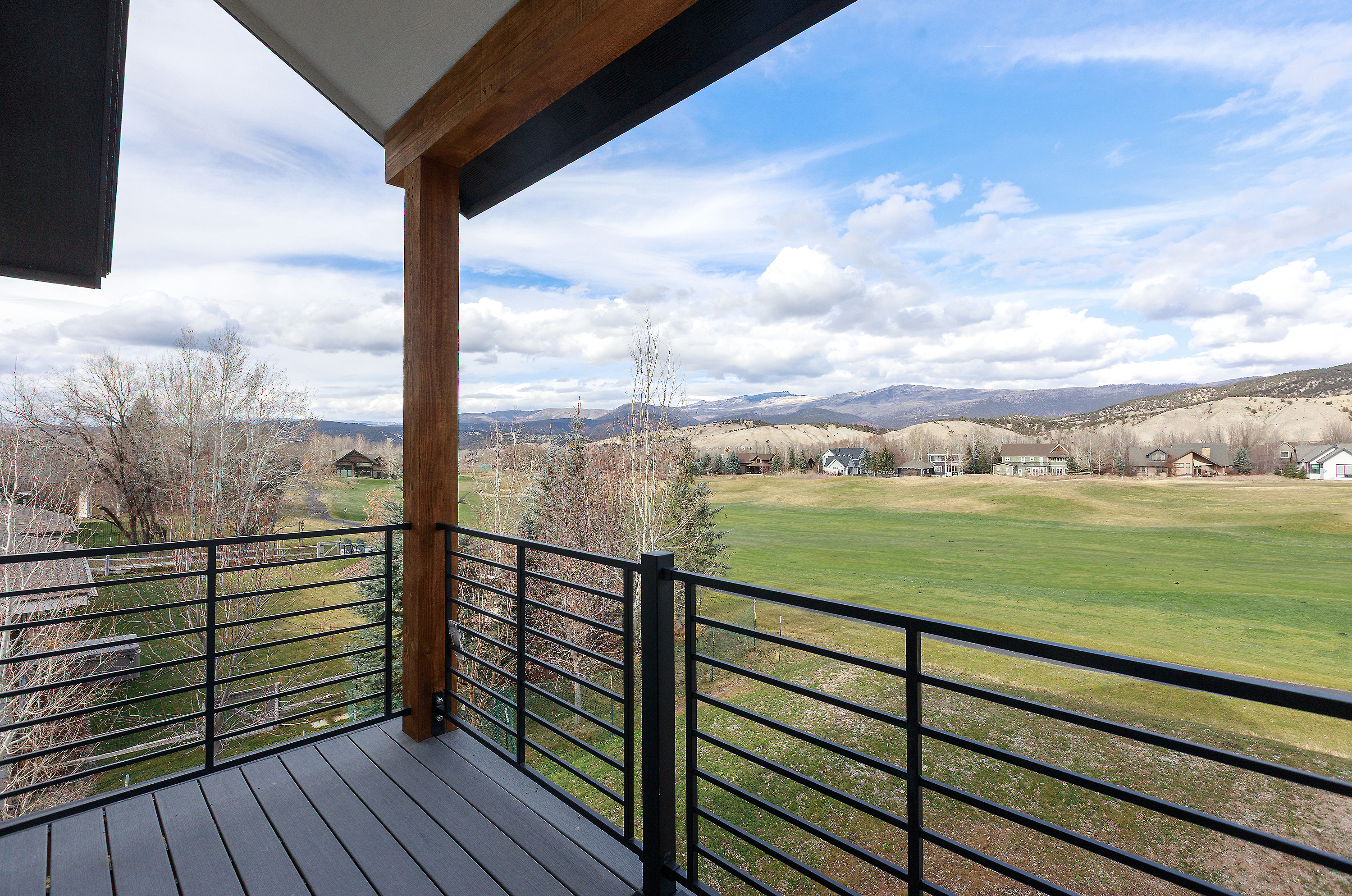 This image showcases a balcony with a modern design, featuring dark gray decking and sleek black metal railings. The view from the balcony overlooks a lush green landscape with scattered houses and distant mountains under a partly cloudy sky. The wooden support beam adds a touch of rustic charm to the contemporary aesthetic.