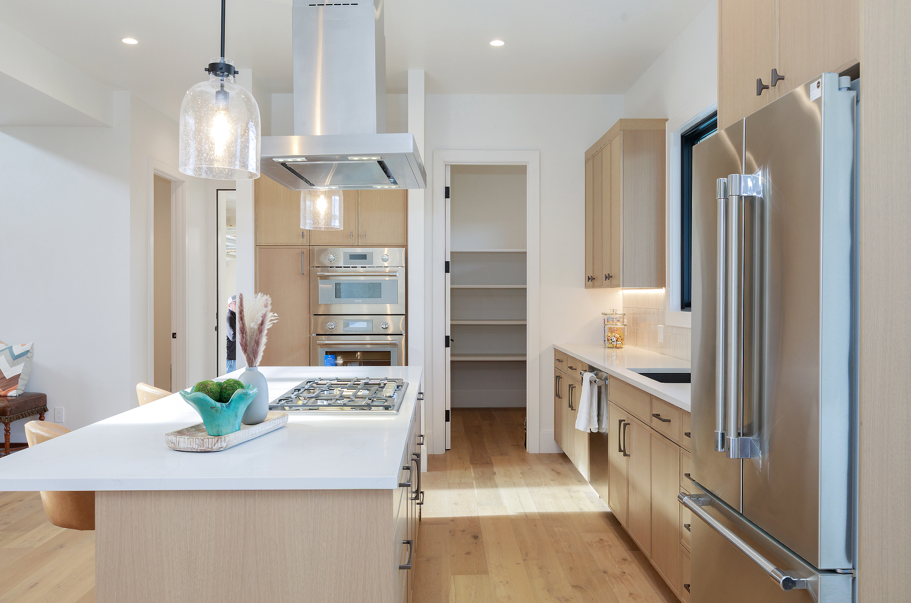 This is a bright and modern kitchen featuring light wood cabinetry and stainless steel appliances, including a large refrigerator and built-in ovens. A central island with a white countertop houses a gas cooktop and provides ample workspace. A walk-in pantry is visible, adding to the kitchen's functionality and storage capacity.