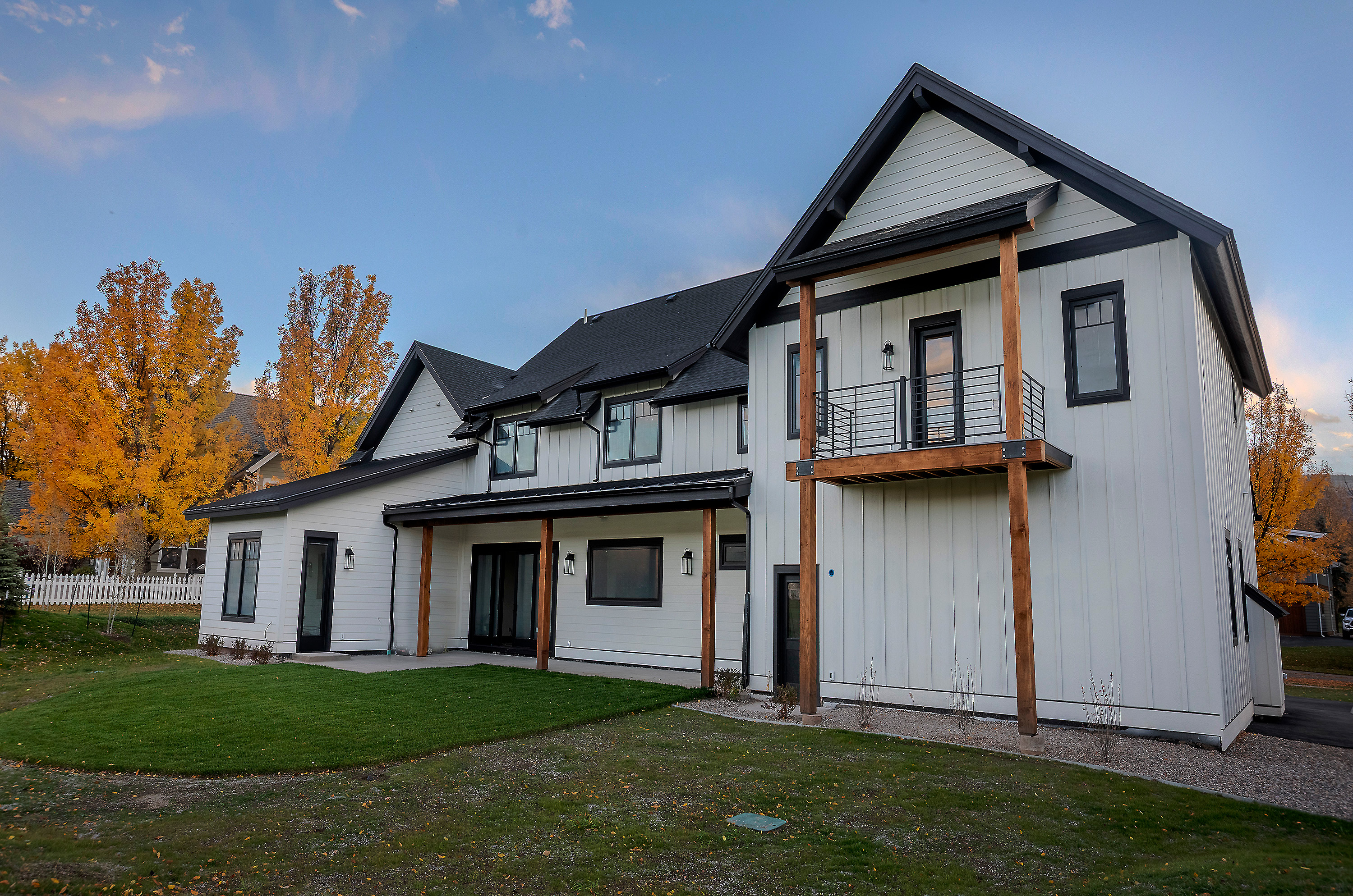 This is a rear view of a modern farmhouse-style home with white siding, black trim, and a dark roof. The house features a second-story balcony with black railings and wooden support beams. The backyard includes a well-maintained lawn and landscaping, creating an inviting outdoor space.