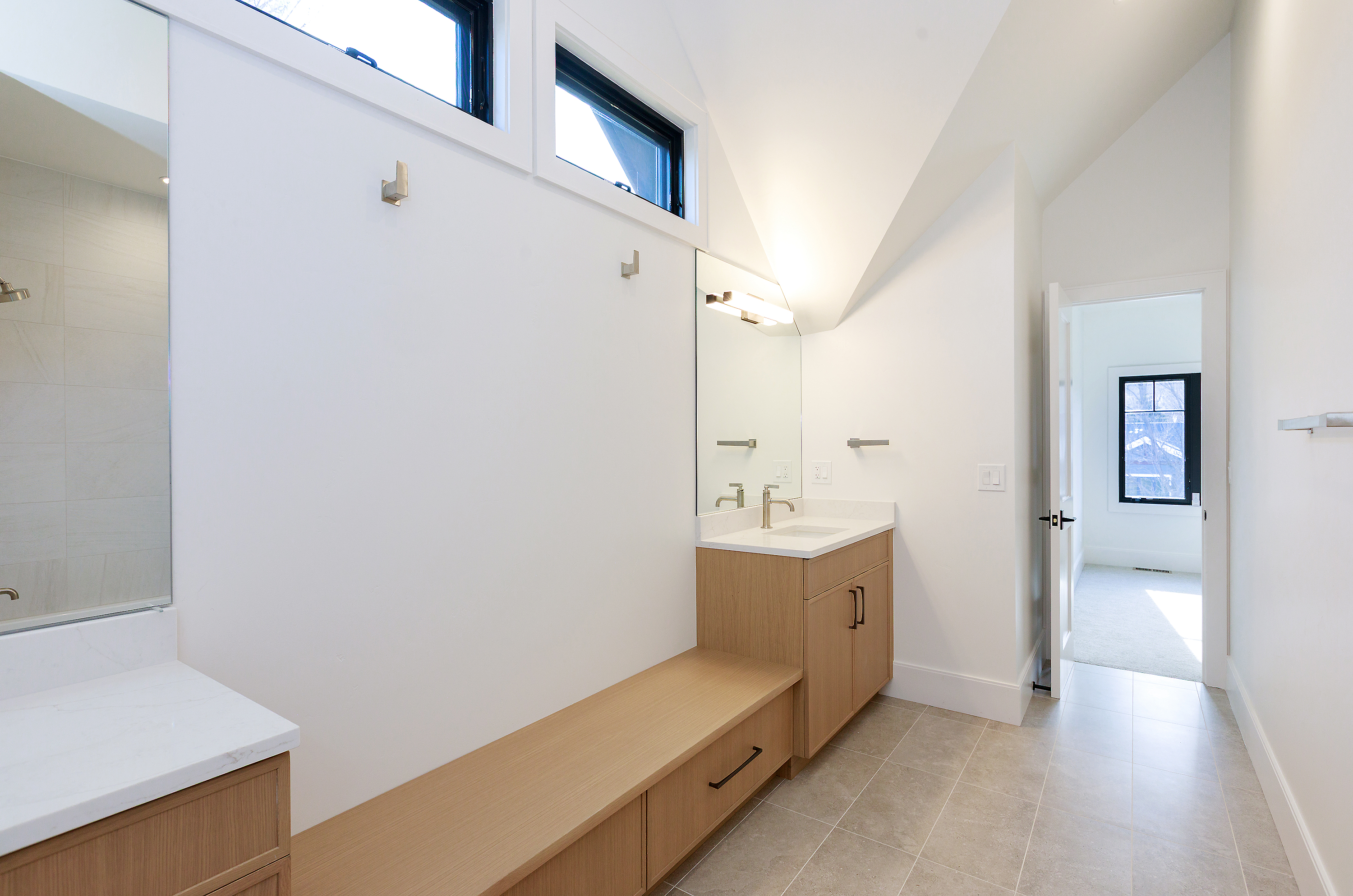 This is a bright and modern primary bathroom featuring a light wood vanity with a white countertop and a large mirror. The room has a clean, minimalist design with white walls, light tile flooring, and clerestory windows that provide natural light. A built-in bench extends from the vanity, offering additional storage and seating.