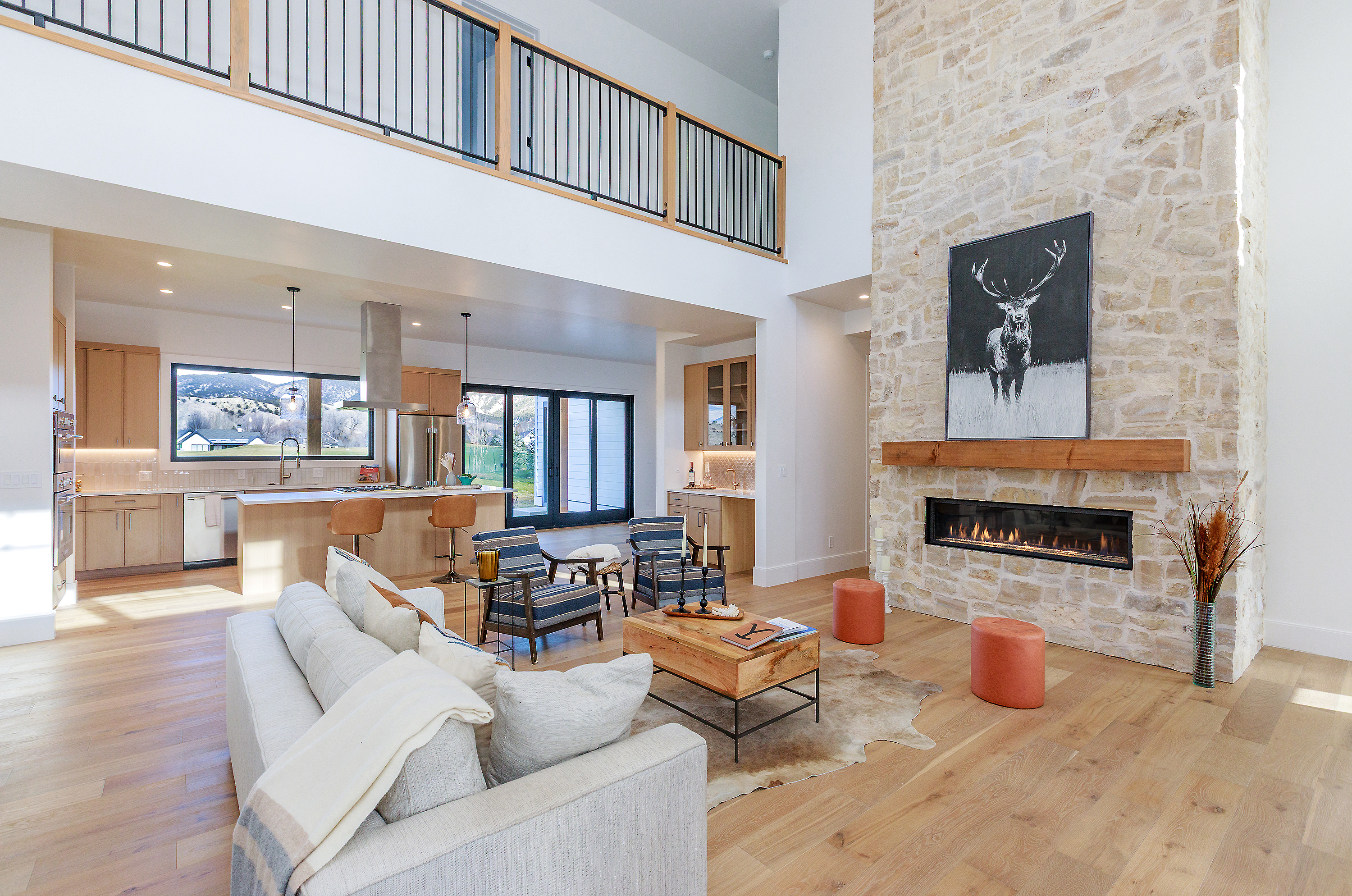 This is a spacious, open-concept living room featuring a stone fireplace with a mounted artwork, hardwood floors, and a comfortable-looking sofa. The room seamlessly connects to the kitchen area, creating a bright and inviting atmosphere. A balcony overlooks the living space, adding architectural interest and a sense of verticality.