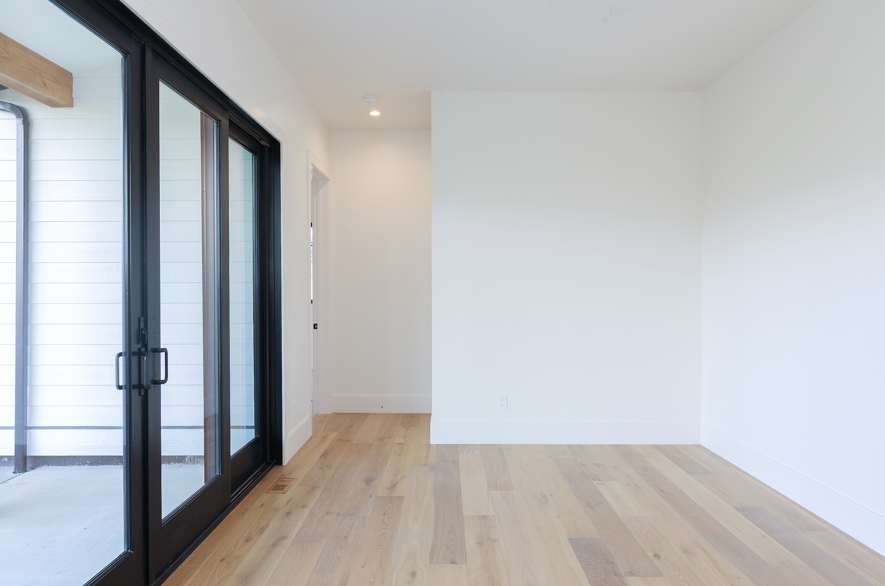 The image showcases a bright and airy living room with light wood flooring and white walls, creating a modern and minimalist aesthetic. A large sliding glass door provides ample natural light and access to an outdoor area. The room appears spacious and clean, ready for personalization.