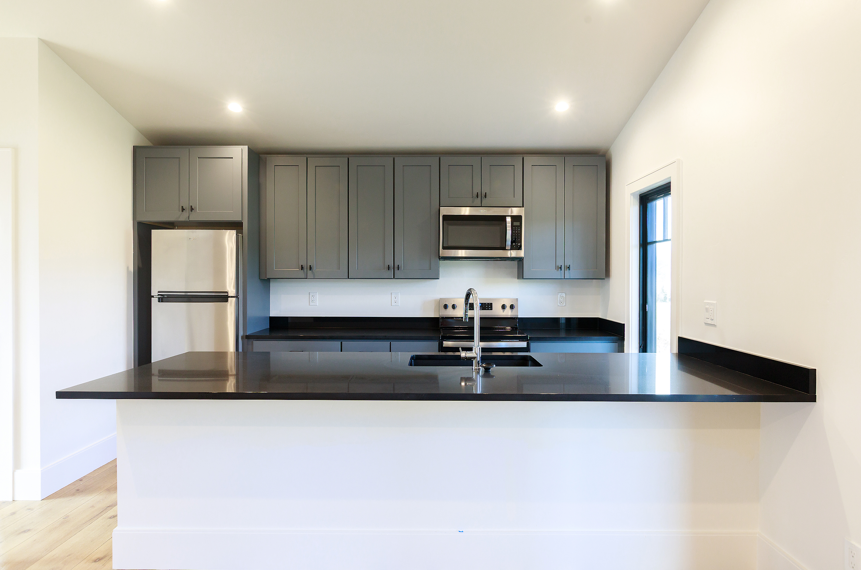 This is a well-lit kitchen featuring gray cabinets, stainless steel appliances, and dark countertops. A kitchen island with a white base provides additional counter space and a sink. The perspective is from the front of the island, showcasing the entire kitchen layout.
