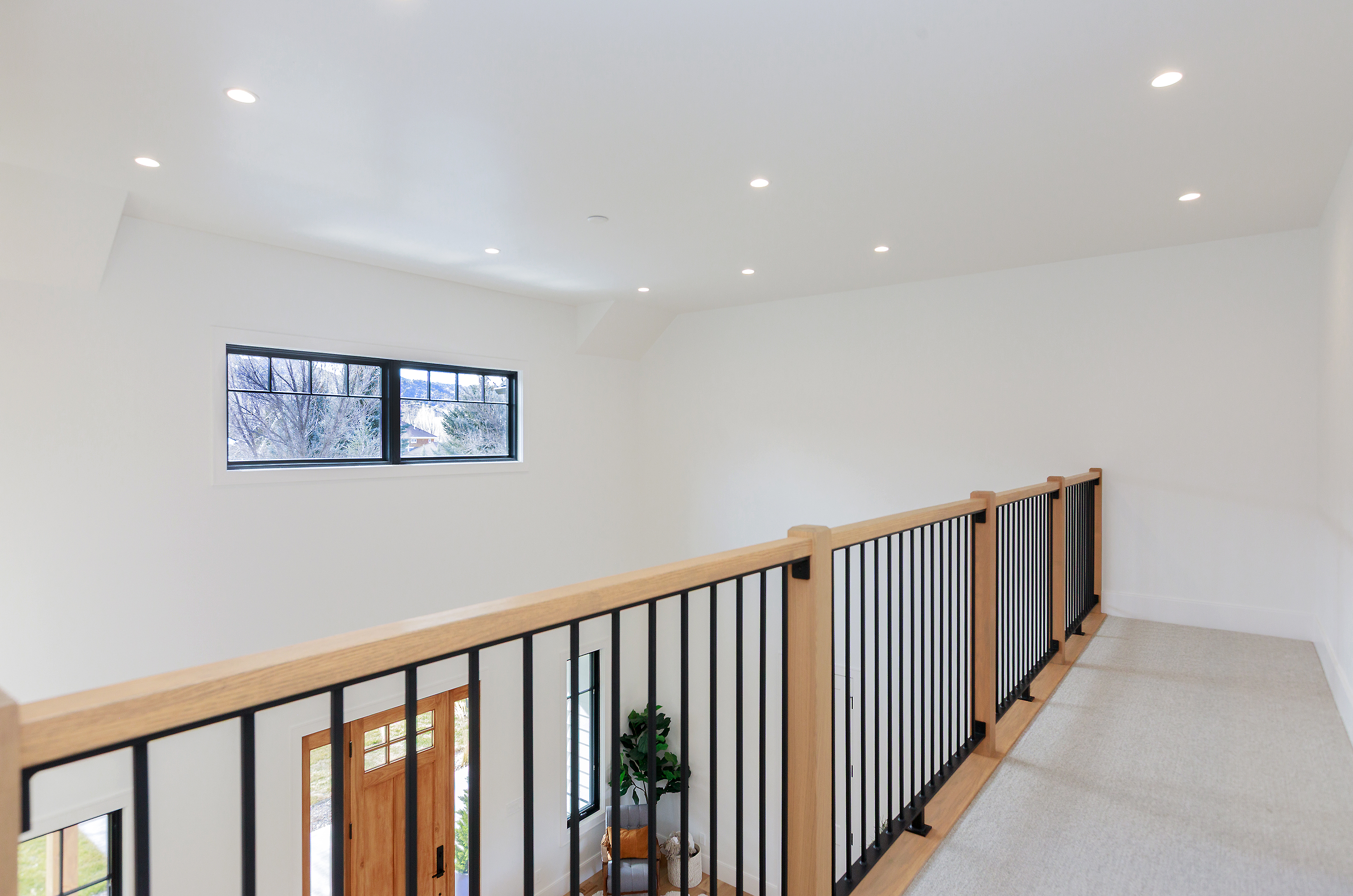 This interior shot showcases a modern hallway with a light-colored carpet and a stylish railing featuring a wooden top and black metal spindles. Natural light streams in through a window, illuminating the white walls and ceiling. The overall impression is clean, bright, and contemporary, highlighting the home's modern design elements.