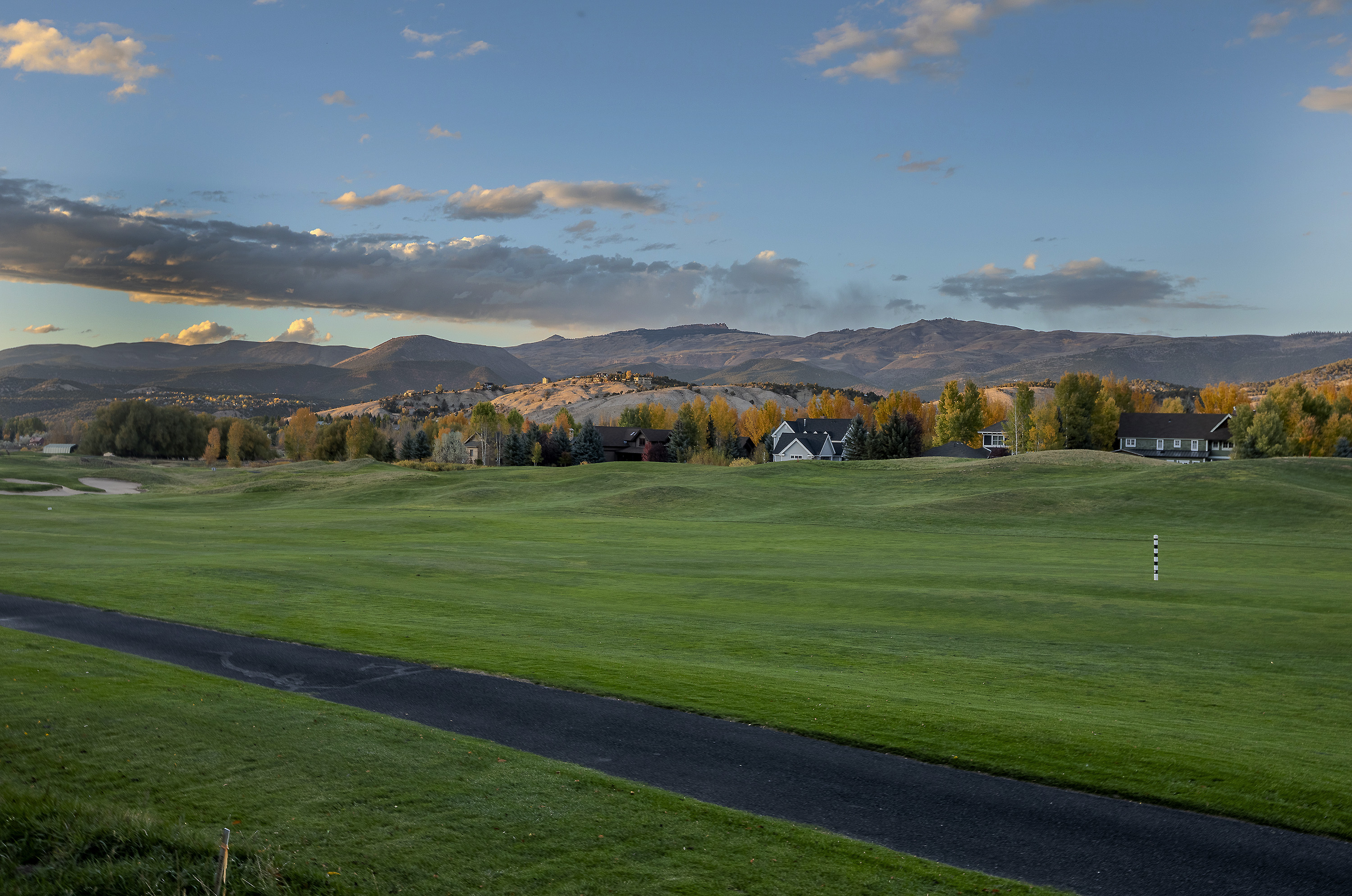 This image showcases a beautifully maintained golf course with several houses in the background, set against a backdrop of rolling mountains under a partly cloudy sky. The lush green fairway is the focal point, with a paved path winding through the landscape, suggesting a peaceful and upscale residential community. The scene evokes a sense of tranquility and luxury, ideal for potential buyers seeking a serene living environment.