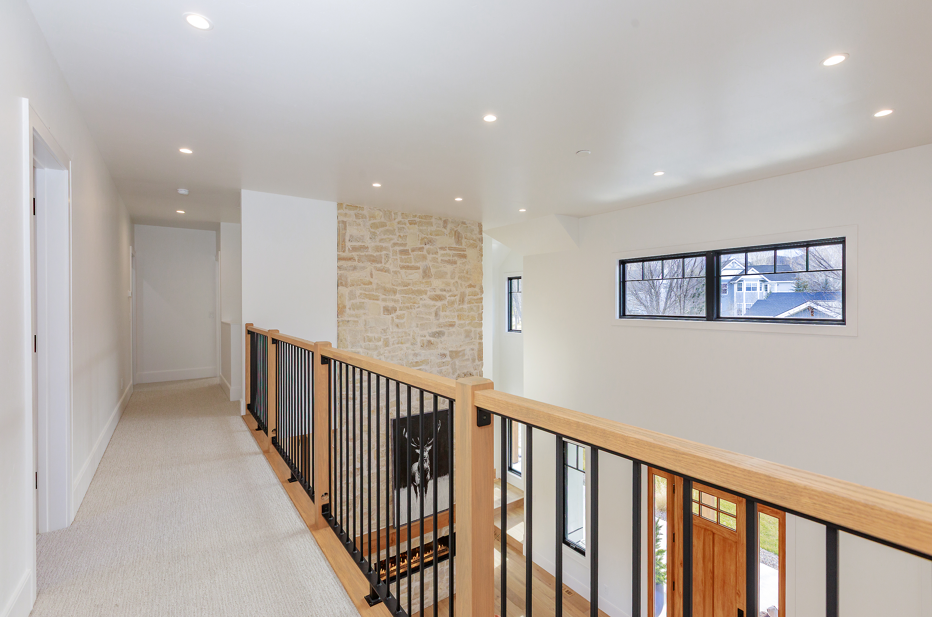 This image showcases a bright and airy hallway with a modern aesthetic. The hallway features a light-colored carpet, white walls, and a wooden and black metal railing. A stone accent wall adds texture and visual interest, while recessed lighting illuminates the space, creating a welcoming and upscale atmosphere.