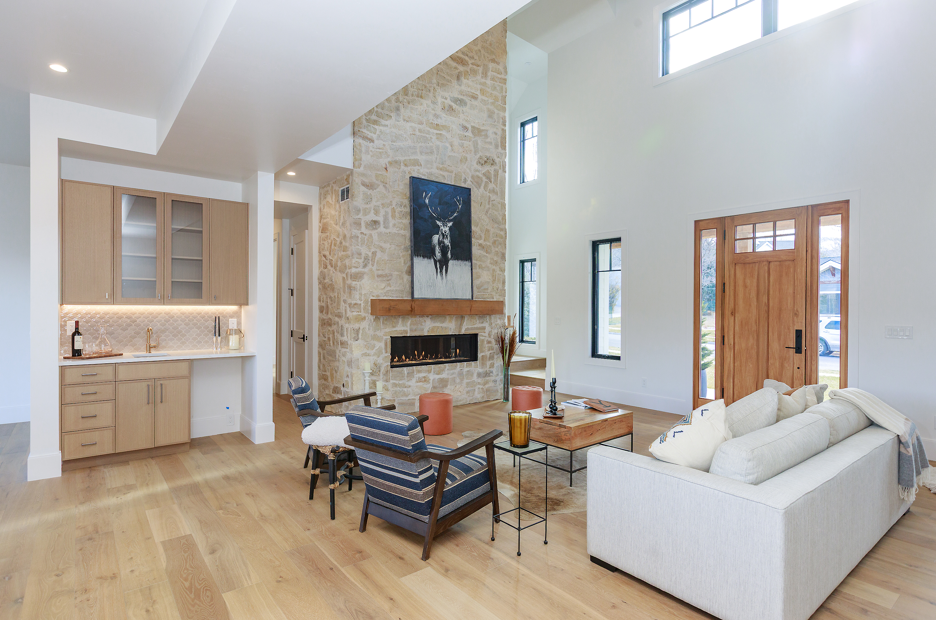 This is an interior shot of a modern living room featuring a stone fireplace with a mounted artwork above. The room is well-lit with natural light coming through large windows, and the decor includes a light-colored sofa, striped armchairs, and a wooden coffee table. A built-in bar area is visible on the left, adding to the room's functionality and appeal.