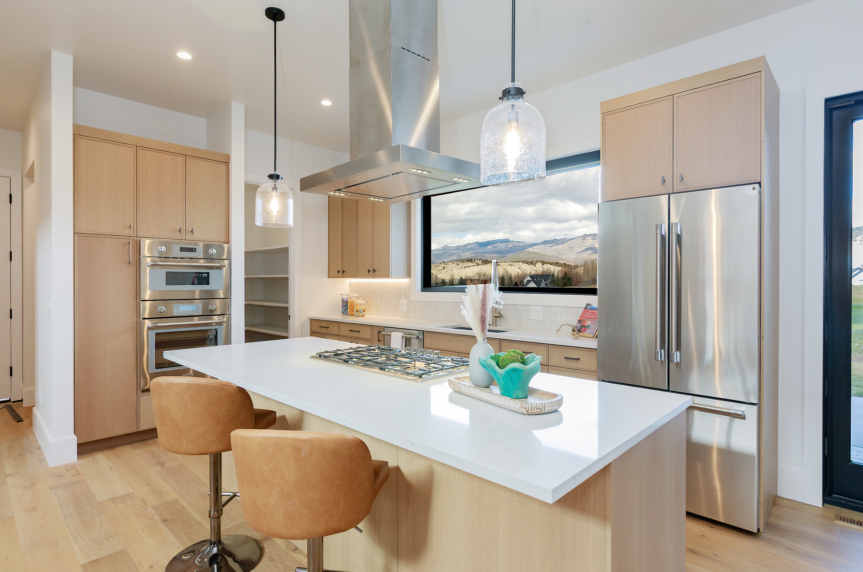 This is a bright and modern kitchen featuring light wood cabinetry, stainless steel appliances, and a large island with a white countertop. Pendant lights hang above the island, and a large window provides a scenic view of the mountains. The kitchen is well-lit and appears spacious, creating an inviting atmosphere.