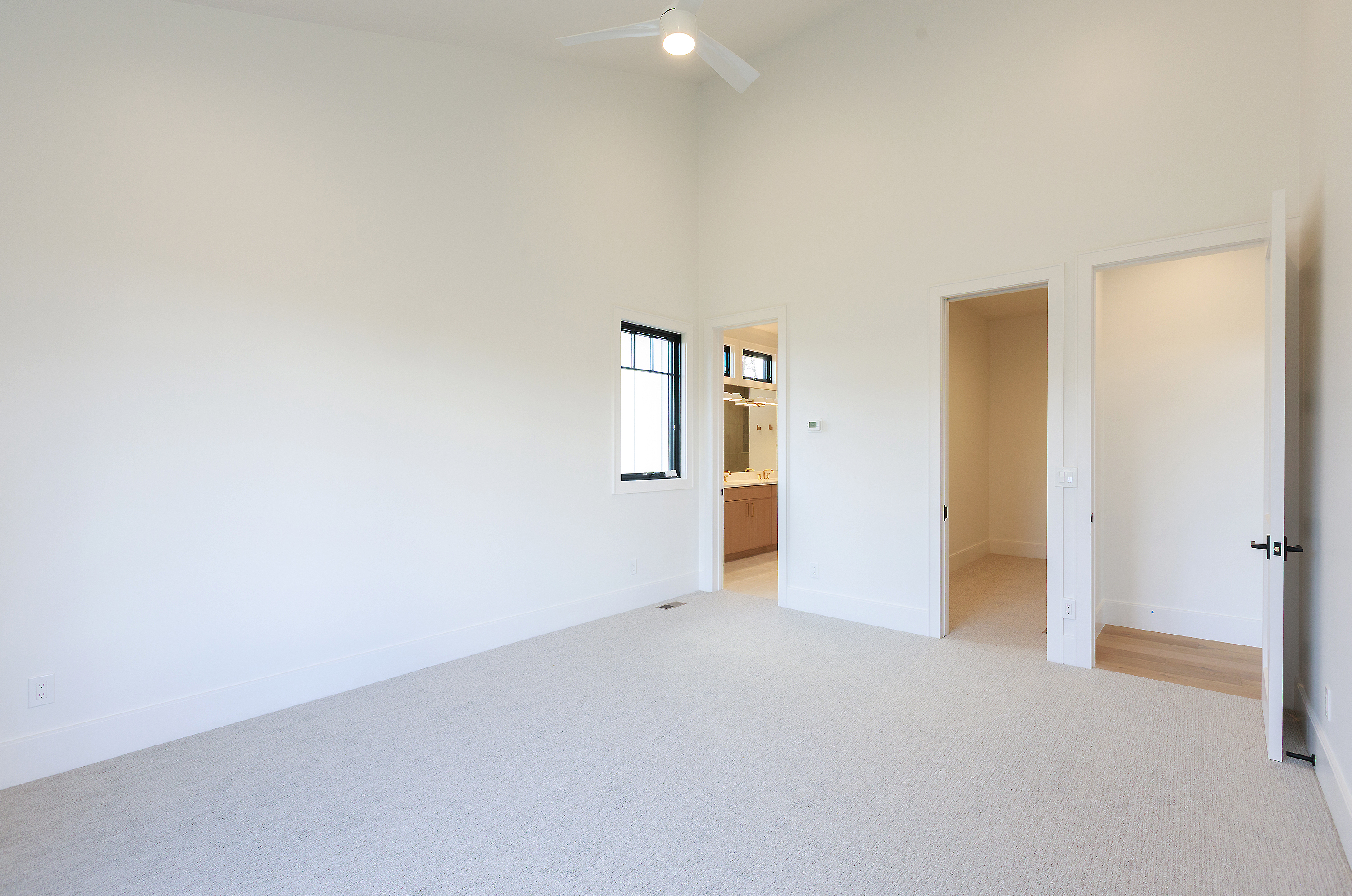 This is a spacious primary bedroom featuring neutral-toned carpet, white walls, and high ceilings, creating a bright and airy atmosphere. A black-framed window provides natural light, while doorways lead to a bathroom and closet, enhancing the room's functionality. The room's minimalist design offers a blank canvas for personalization.