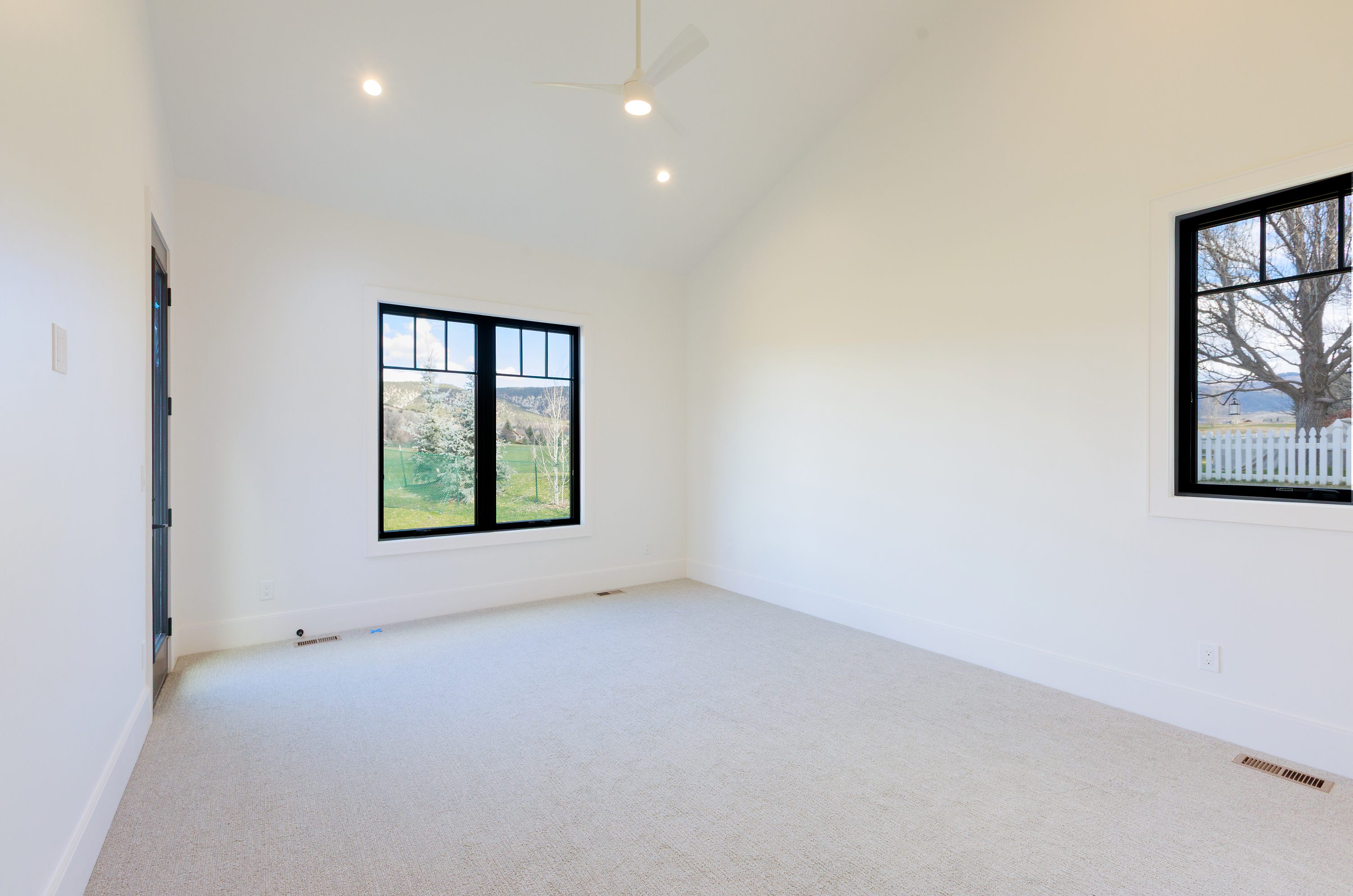 This is a bright and spacious primary bedroom featuring neutral carpet, white walls, and black-framed windows that offer views of the surrounding landscape. The room has a minimalist aesthetic with clean lines and a vaulted ceiling, creating an airy and inviting atmosphere. A ceiling fan provides air circulation.
