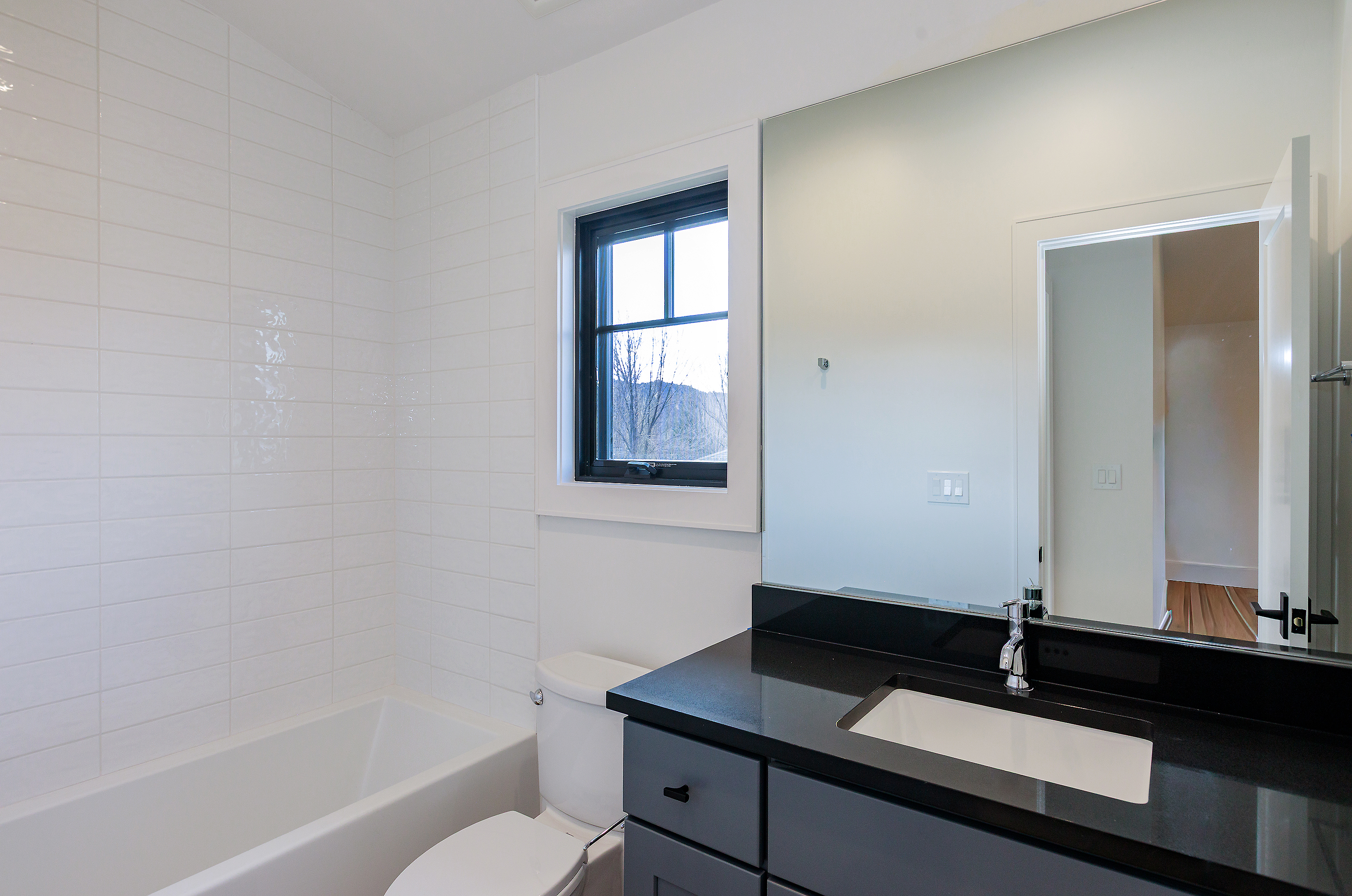 This is a well-lit bathroom featuring a white bathtub with white tiled walls, a toilet, and a vanity with a black countertop and gray cabinets. A black-framed window provides natural light. The mirror reflects an open doorway, adding depth to the space and showcasing the clean, modern design.