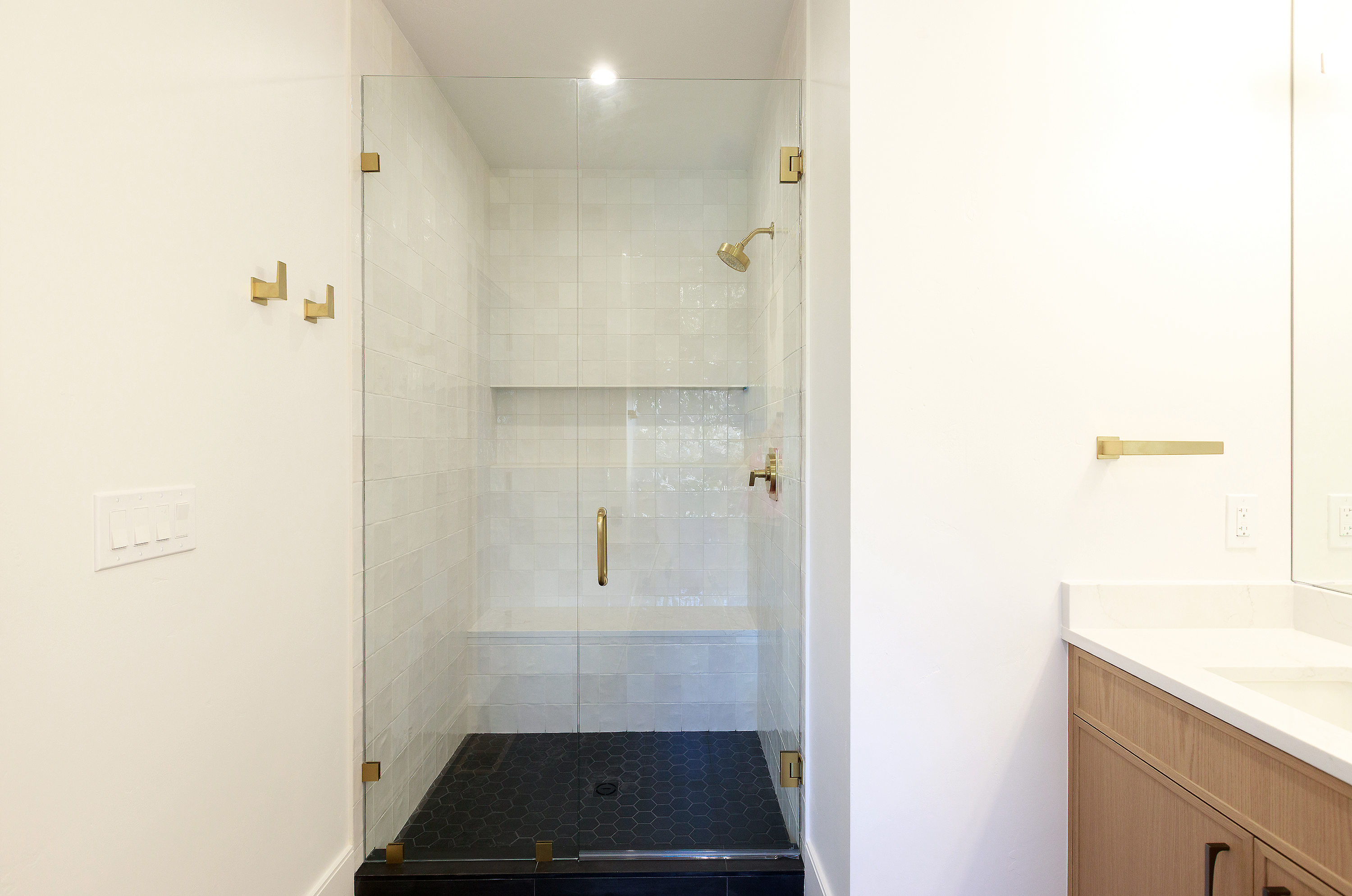 This is a bright and modern bathroom featuring a glass-enclosed shower with white tiled walls and a dark, hexagonal tiled floor. The shower has gold fixtures, including the shower head and handle, adding a touch of elegance. A light wood vanity with a white countertop is visible on the right, complementing the clean and minimalist design.