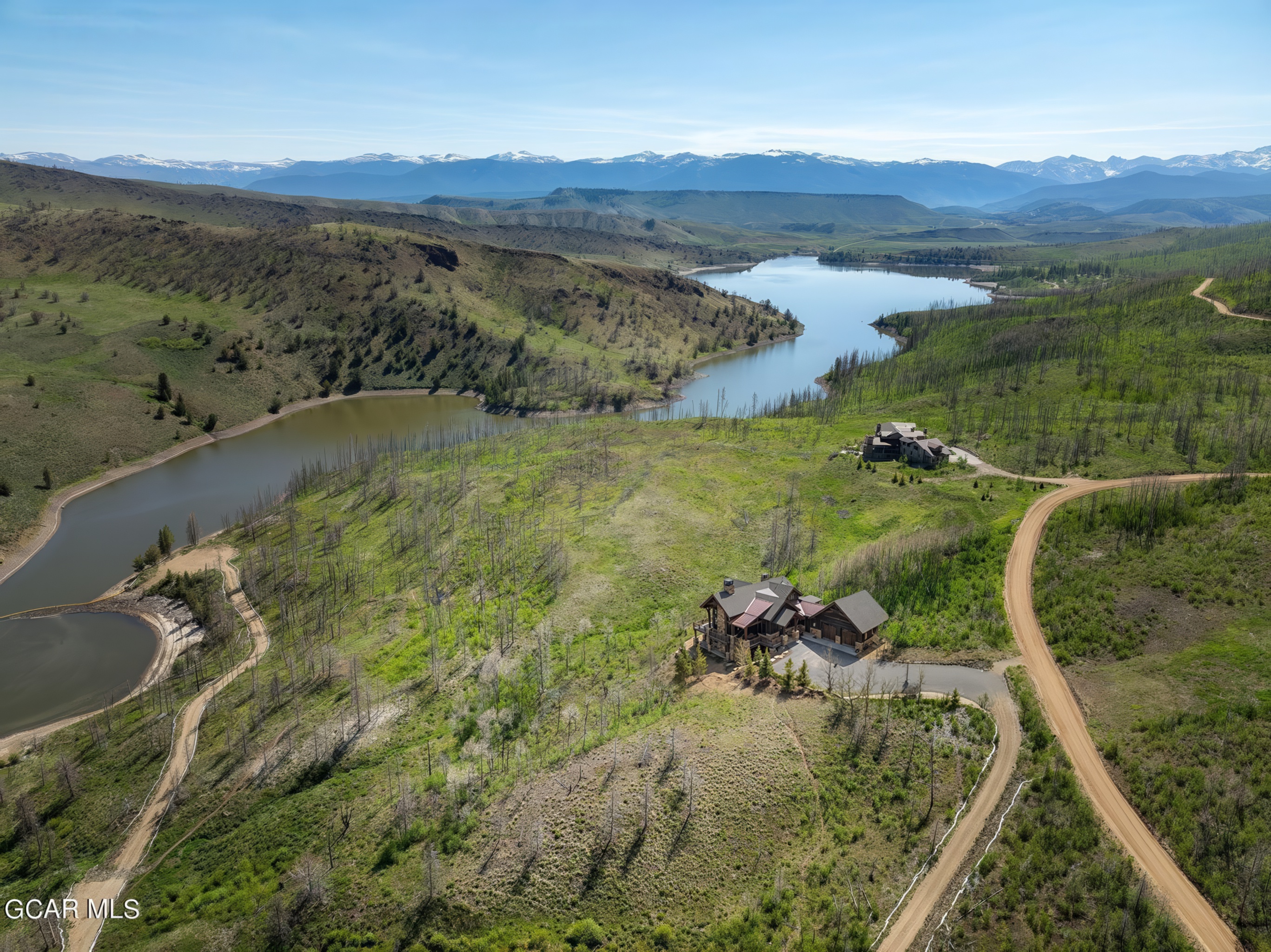 This high-altitude aerial perspective captures a sprawling mountain estate nestled within a vast, rugged landscape overlooking a serene lake. The property features a large, multi-gabled residence with a rustic aesthetic, positioned on a grassy hillside surrounded by sparse, weathered trees and winding dirt access roads. In the distance, a majestic range of snow-capped mountains creates a dramatic backdrop, emphasizing the secluded and expansive nature of this rural retreat.