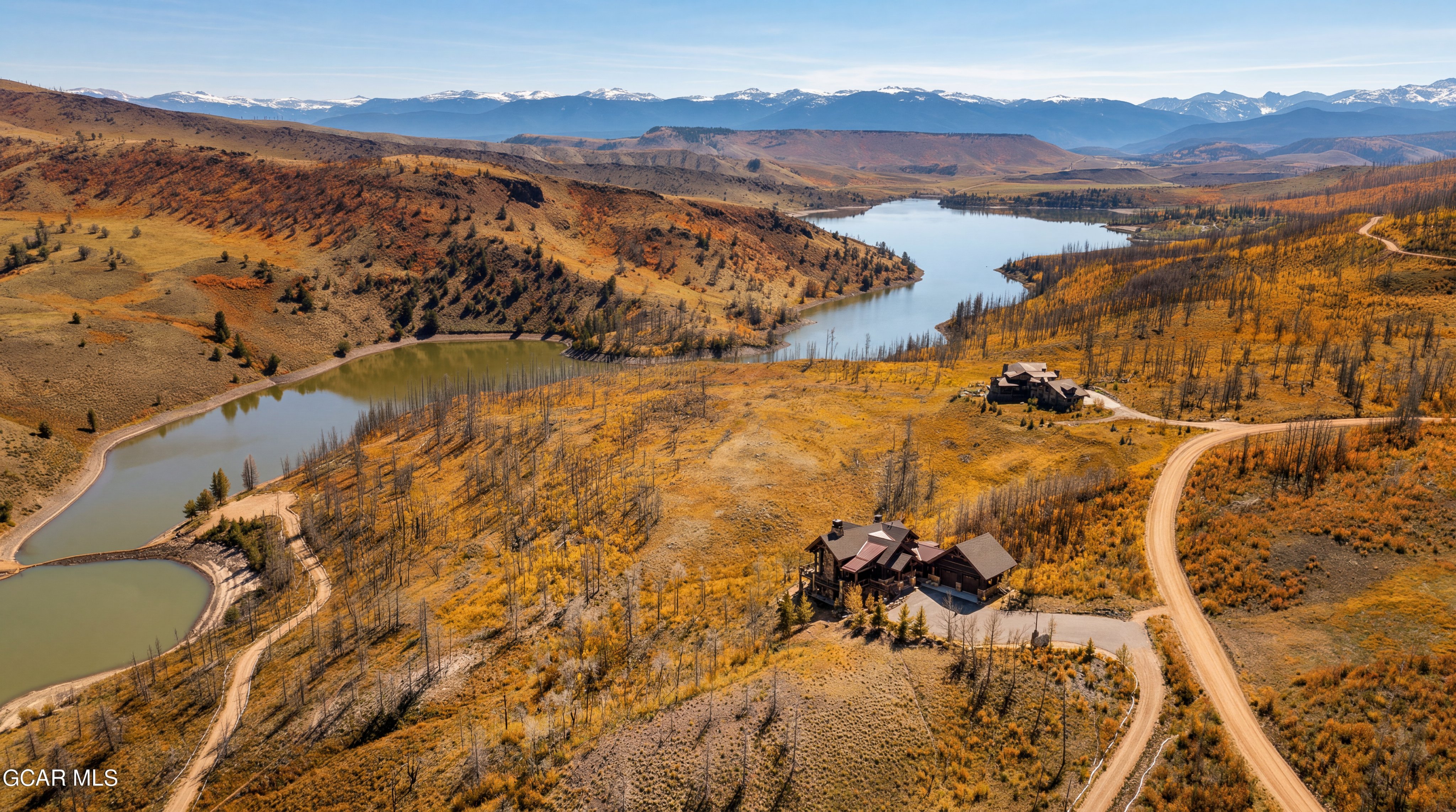 This expansive aerial view captures a secluded mountain estate nestled within a vast, rugged landscape of golden autumn foliage and sparse, weathered trees. The property features a large, multi-gabled residence with a dark roof and stone accents, positioned prominently on a hillside overlooking a serene, winding lake. In the distance, majestic snow-capped mountain peaks frame the horizon, creating a dramatic and picturesque setting for this remote retreat.