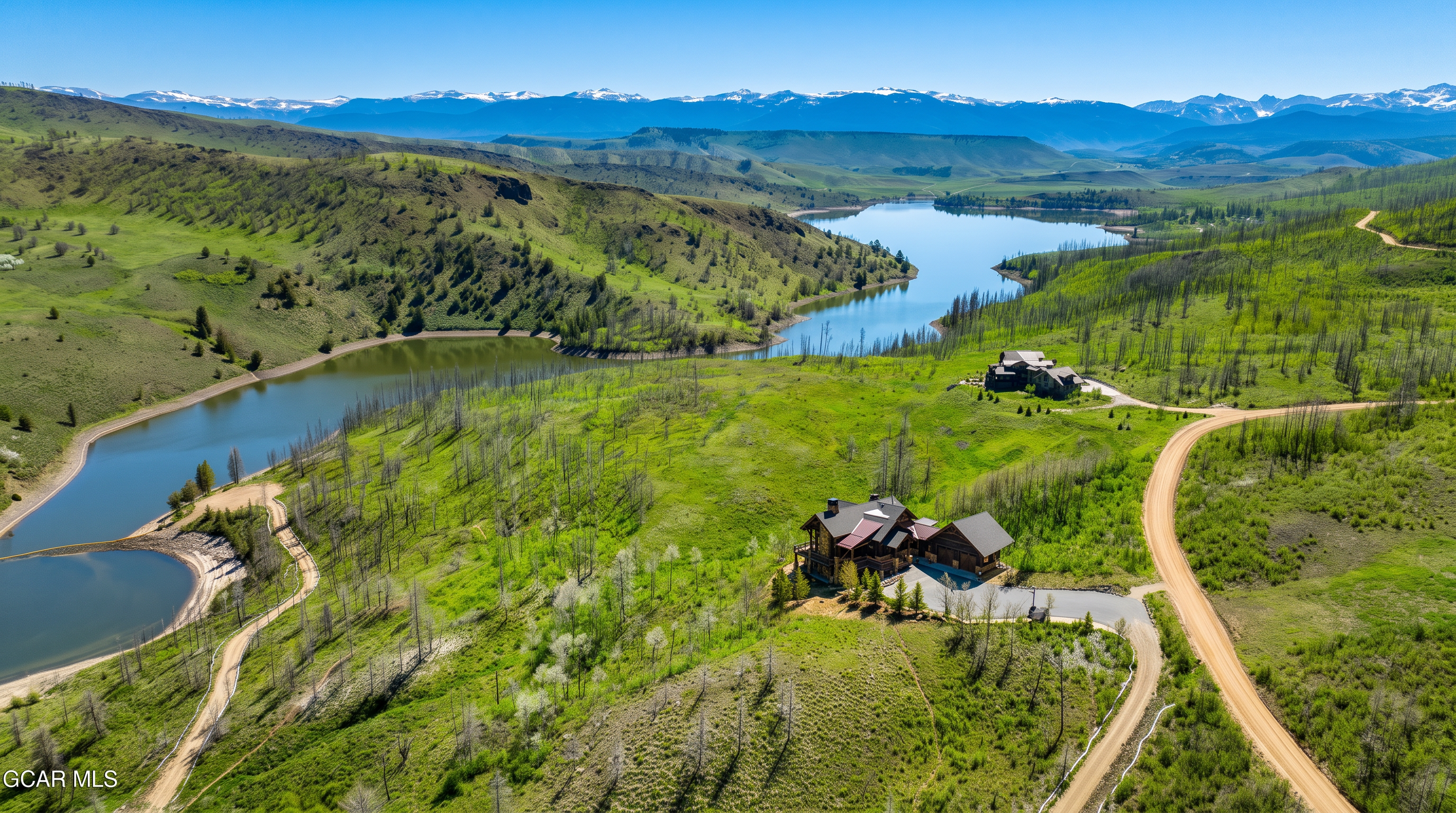 This expansive aerial view captures a secluded mountain estate nestled within a lush, rolling landscape overlooking a serene lake. The property features a large, multi-level residence with a dark-toned roof and wood-accented exterior, positioned prominently on a grassy hillside. A winding dirt road leads to the home, while the backdrop showcases majestic, snow-capped mountain ranges under a clear blue sky.