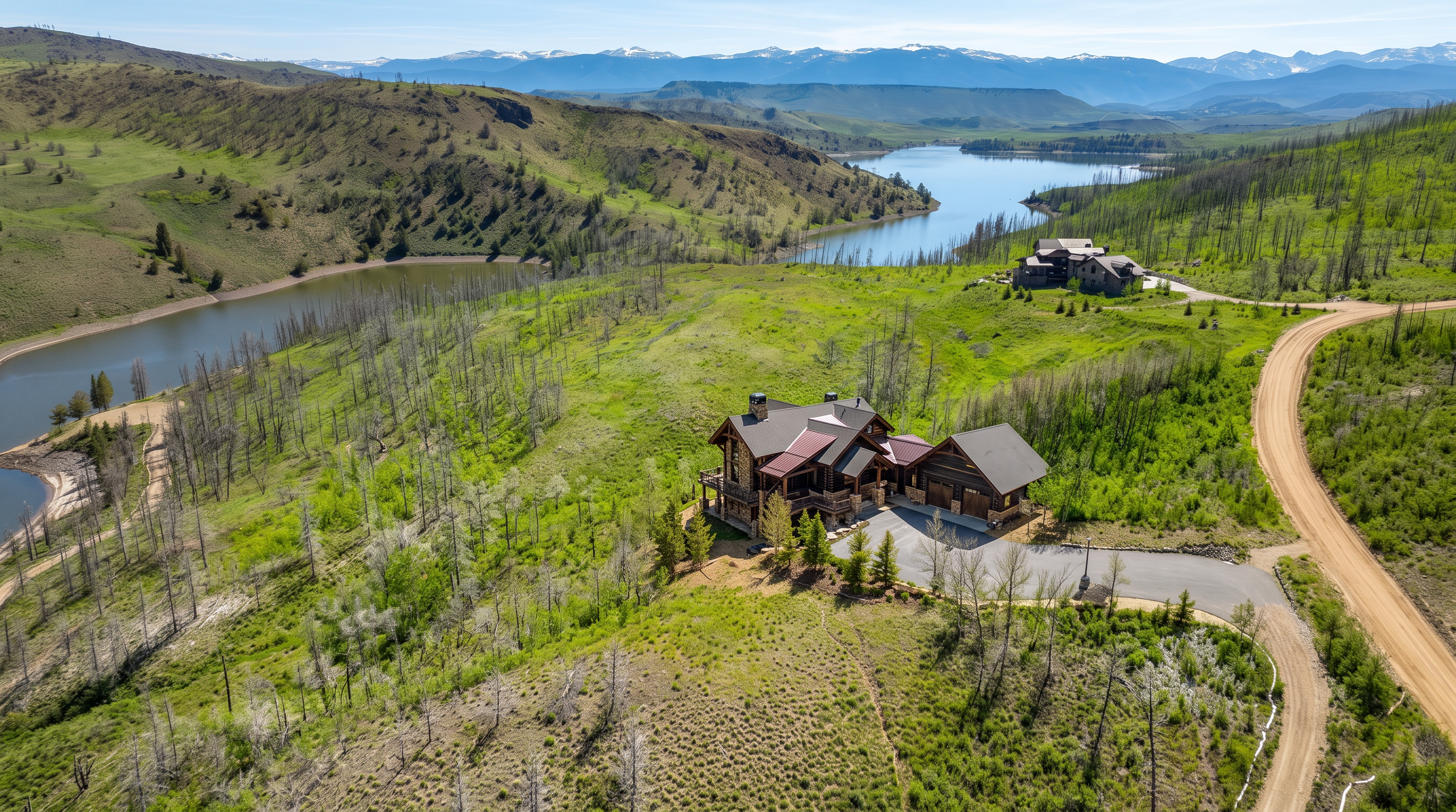 This high-angle aerial perspective captures a sprawling mountain estate nestled within a vast, rugged landscape of rolling green hills and sparse, weathered trees. The primary residence features a rustic architectural style with dark wood siding, a multi-gabled roof with metal accents, and a paved driveway leading to an attached garage. In the distance, a serene lake winds through the valley, framed by majestic, snow-capped mountain peaks under a clear blue sky.
