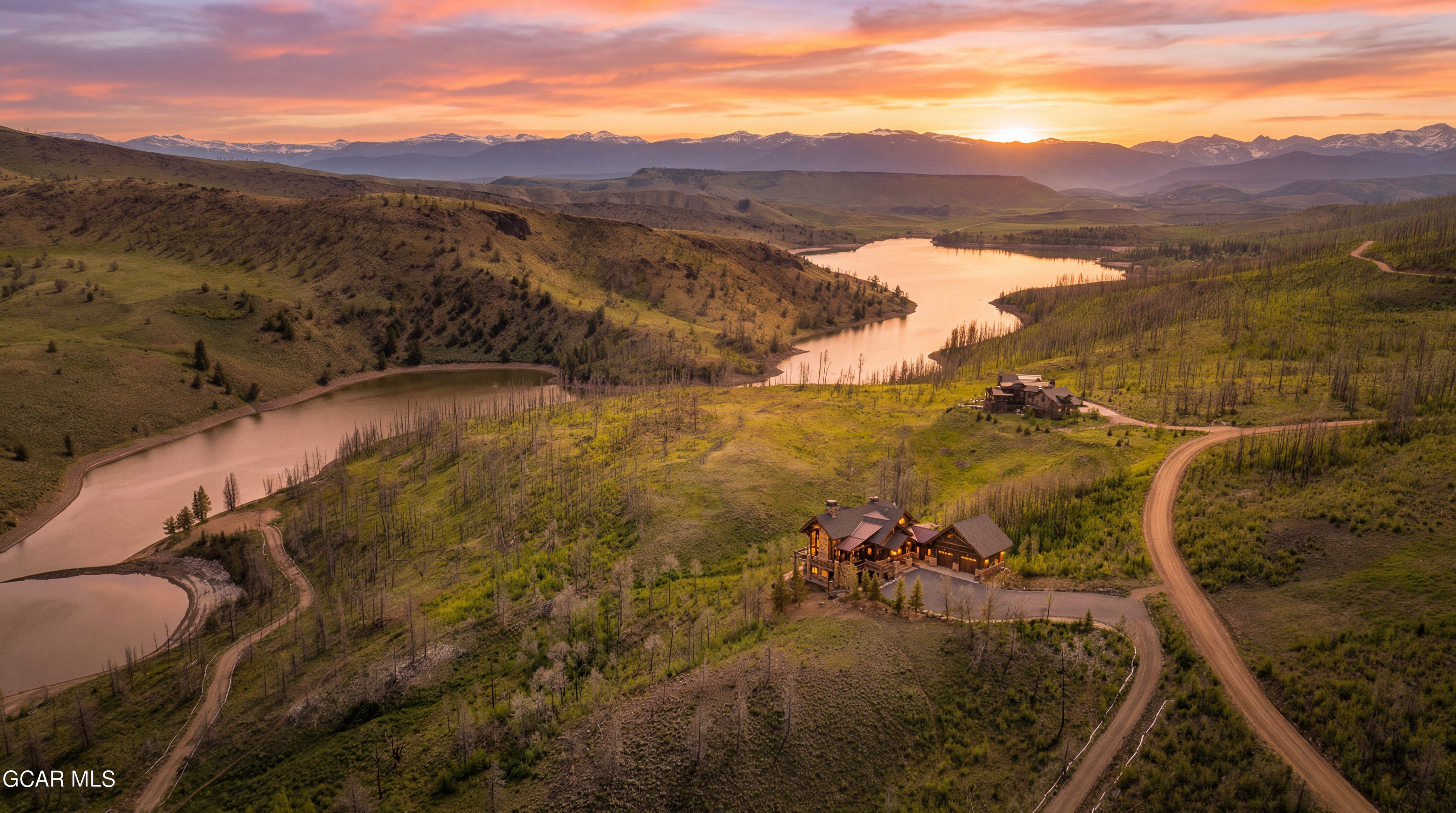 This stunning aerial perspective captures a luxury mountain estate nestled within a vast, rolling landscape at sunset. The property features a large, multi-gabled residence with warm, inviting interior lighting, positioned prominently on a hillside overlooking a serene lake and distant, snow-capped mountain ranges. A winding dirt road leads up to the home, emphasizing the secluded and expansive nature of this scenic, high-altitude retreat.