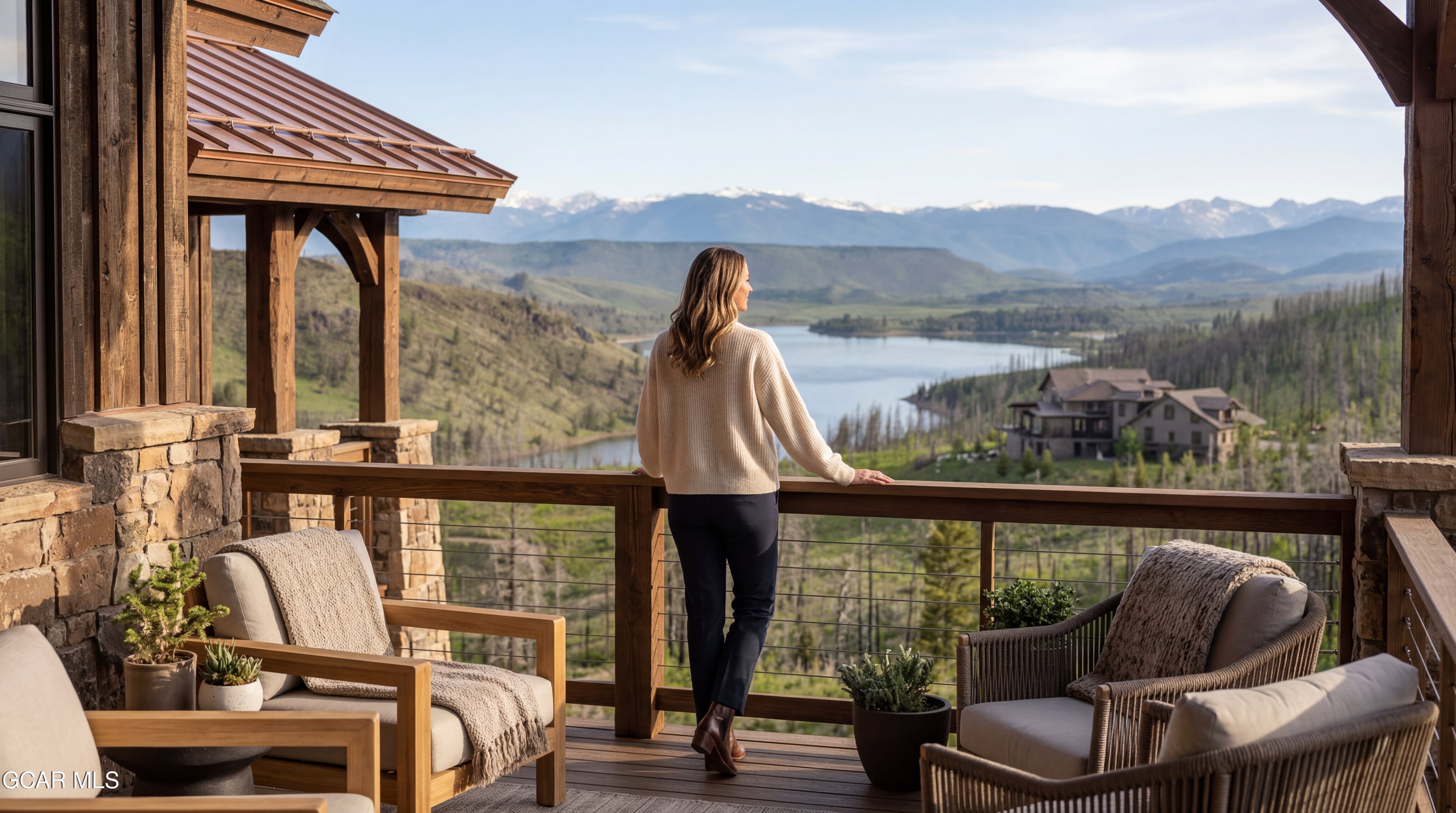 This elevated deck offers a breathtaking panoramic view of a serene lake nestled among rolling hills and distant snow-capped mountains. The space is furnished with comfortable outdoor seating, including wooden armchairs and woven chairs, creating a peaceful retreat for enjoying the scenery. A person stands at the wooden railing, taking in the expansive landscape, which highlights the property's prime location and connection to nature.
