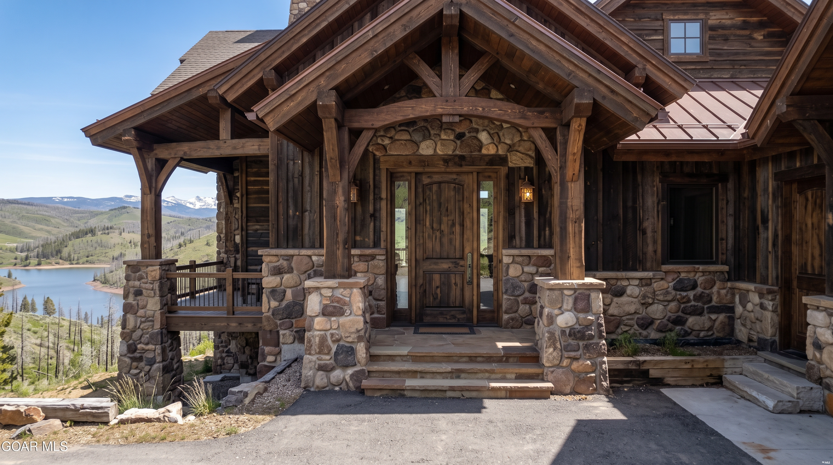 This rustic mountain-style home features a prominent timber-framed entryway with a stone-clad base and a covered porch. The facade is constructed from dark vertical wood siding and natural stone, creating a warm, rugged aesthetic that complements the surrounding landscape. A scenic view of a lake and distant snow-capped mountains is visible from the side deck, emphasizing the property's secluded and picturesque location.