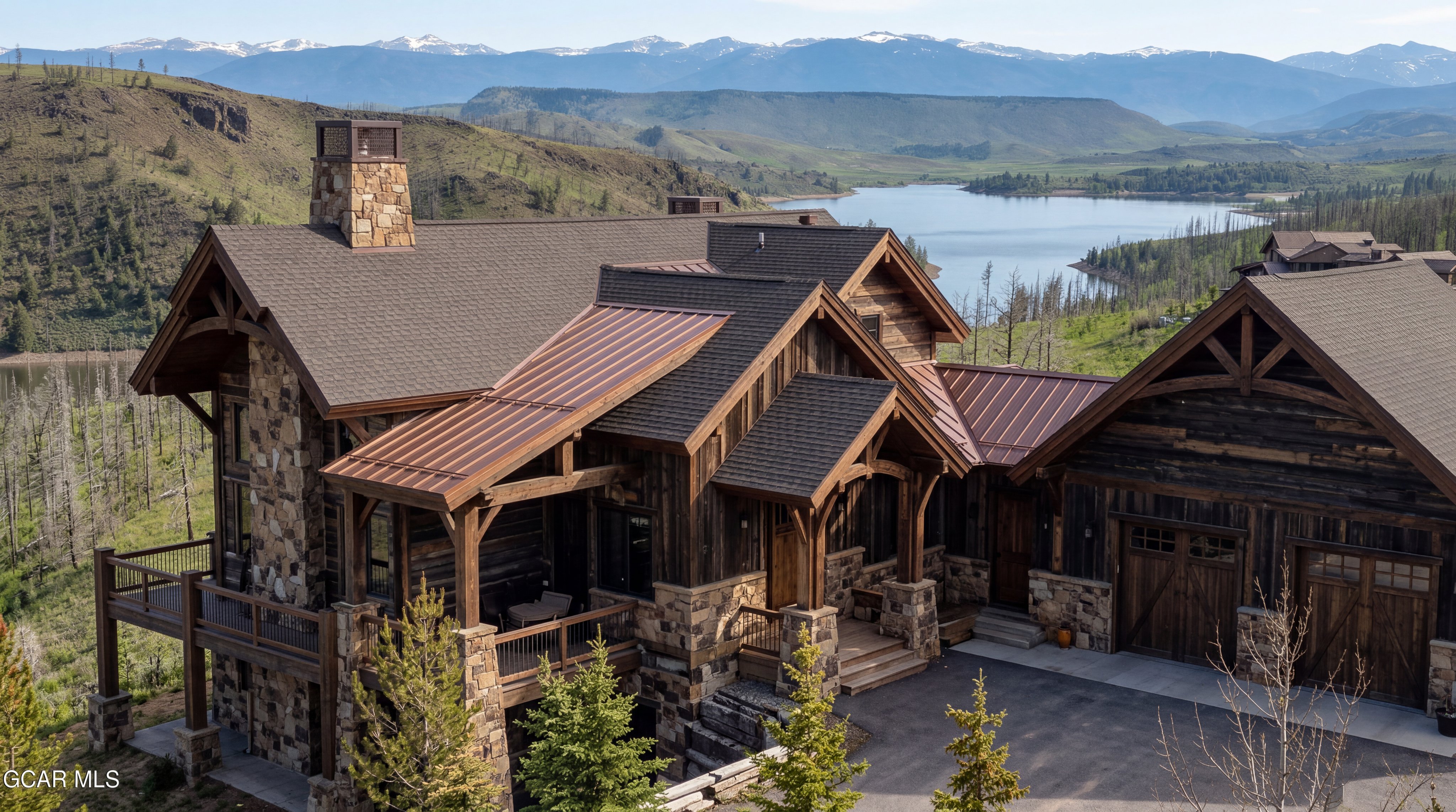 This high-angle aerial view captures a luxurious mountain lodge featuring a blend of rustic timber siding and extensive stone masonry. The home is topped with a combination of dark shingle roofing and striking copper-toned metal accents, while a spacious deck overlooks a serene lake and distant snow-capped peaks. The property is nestled in a rugged, natural landscape, emphasizing its secluded and scenic location.