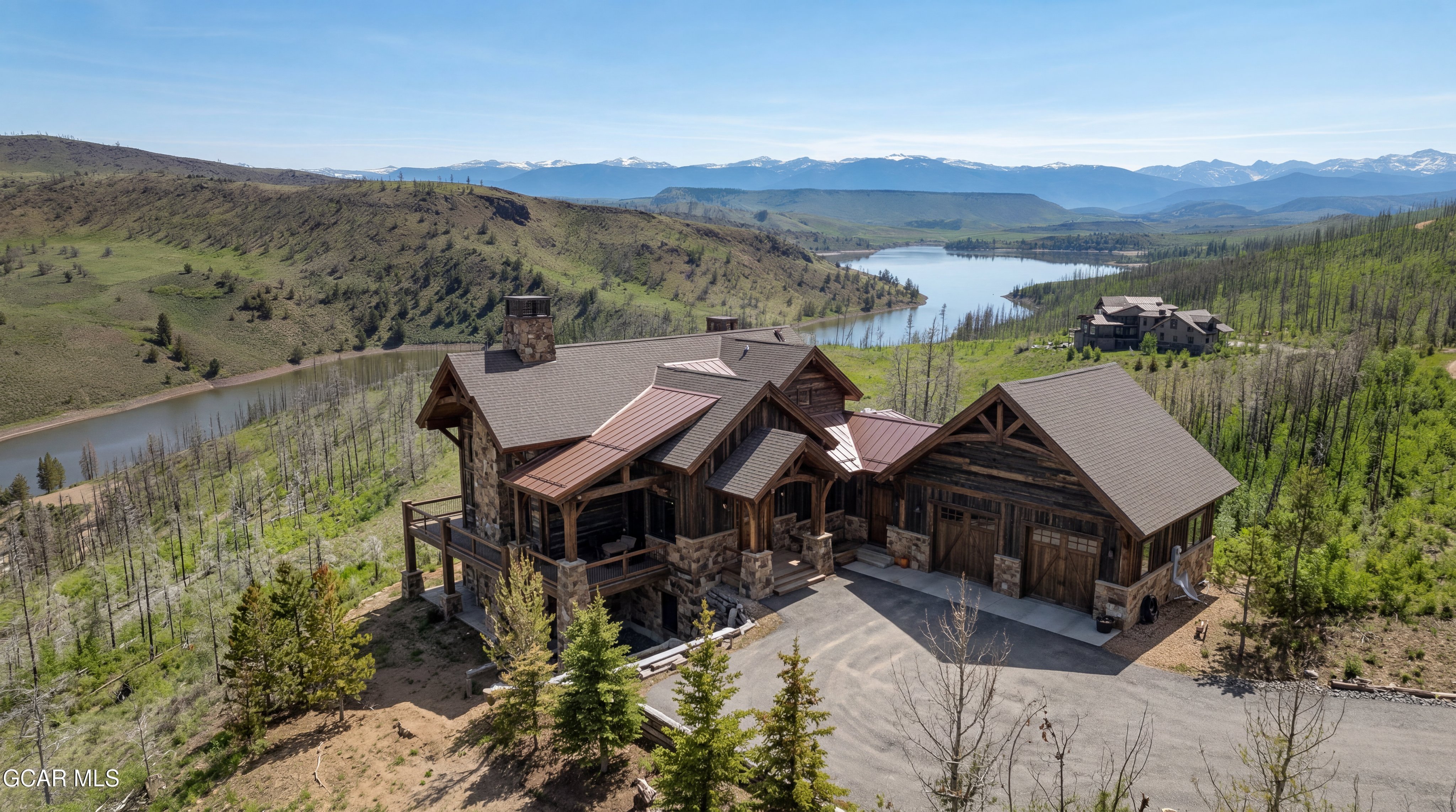 This aerial perspective captures a sprawling mountain lodge set against a dramatic backdrop of a lake and snow-capped peaks. The home features a rustic aesthetic with a combination of dark wood siding, stone accents, and a multi-gabled roofline with metal roofing sections. A paved driveway leads to a two-car garage, while a covered deck overlooks the rugged, forested landscape.