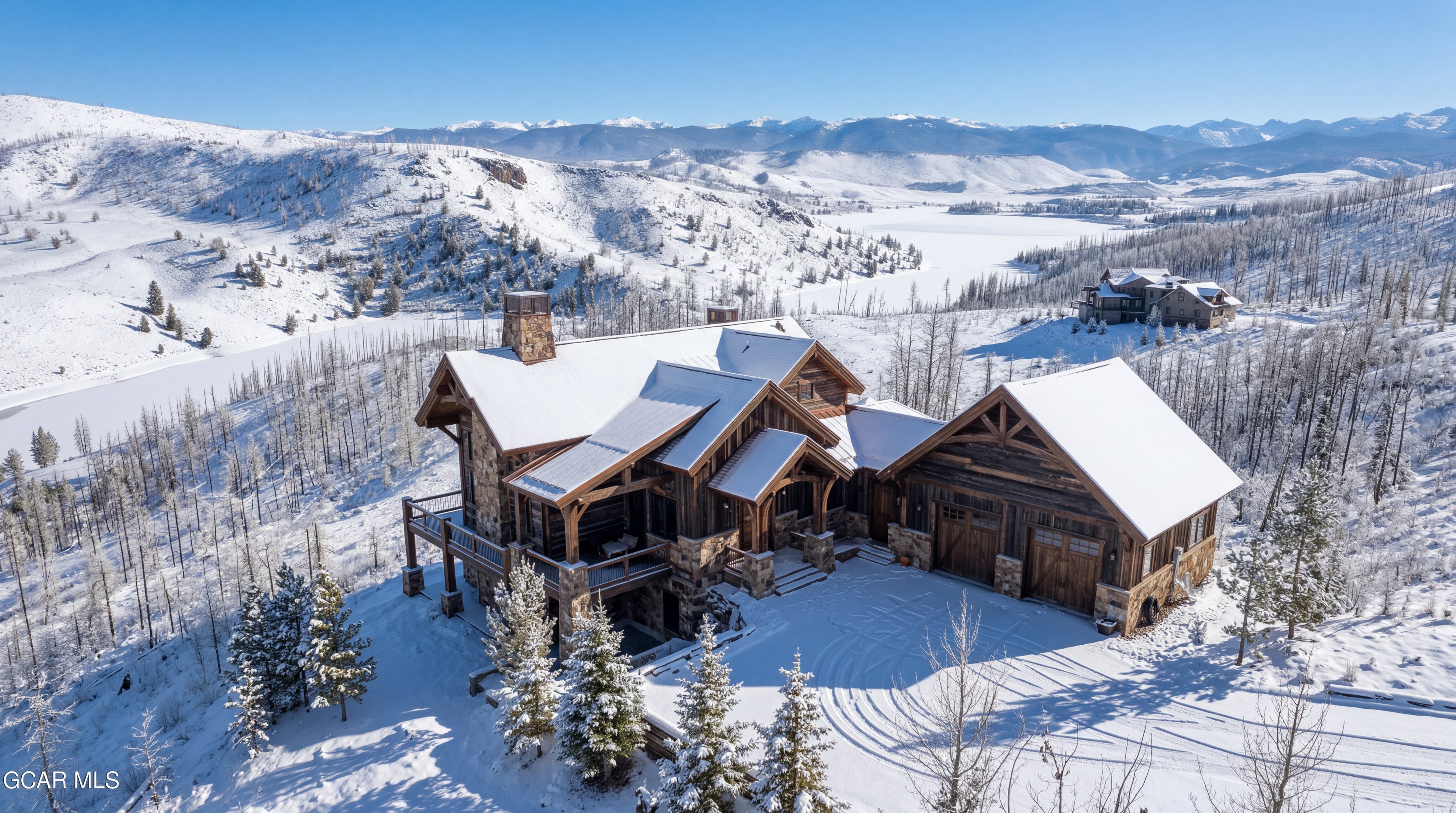This stunning aerial perspective captures a rustic mountain lodge nestled in a snow-covered landscape with a frozen lake in the distance. The home features a multi-gabled roofline, natural wood siding, and stone accents, complemented by a spacious deck and an attached garage. The surrounding terrain is filled with evergreen trees and rolling, snow-dusted hills, creating a serene and secluded alpine atmosphere.