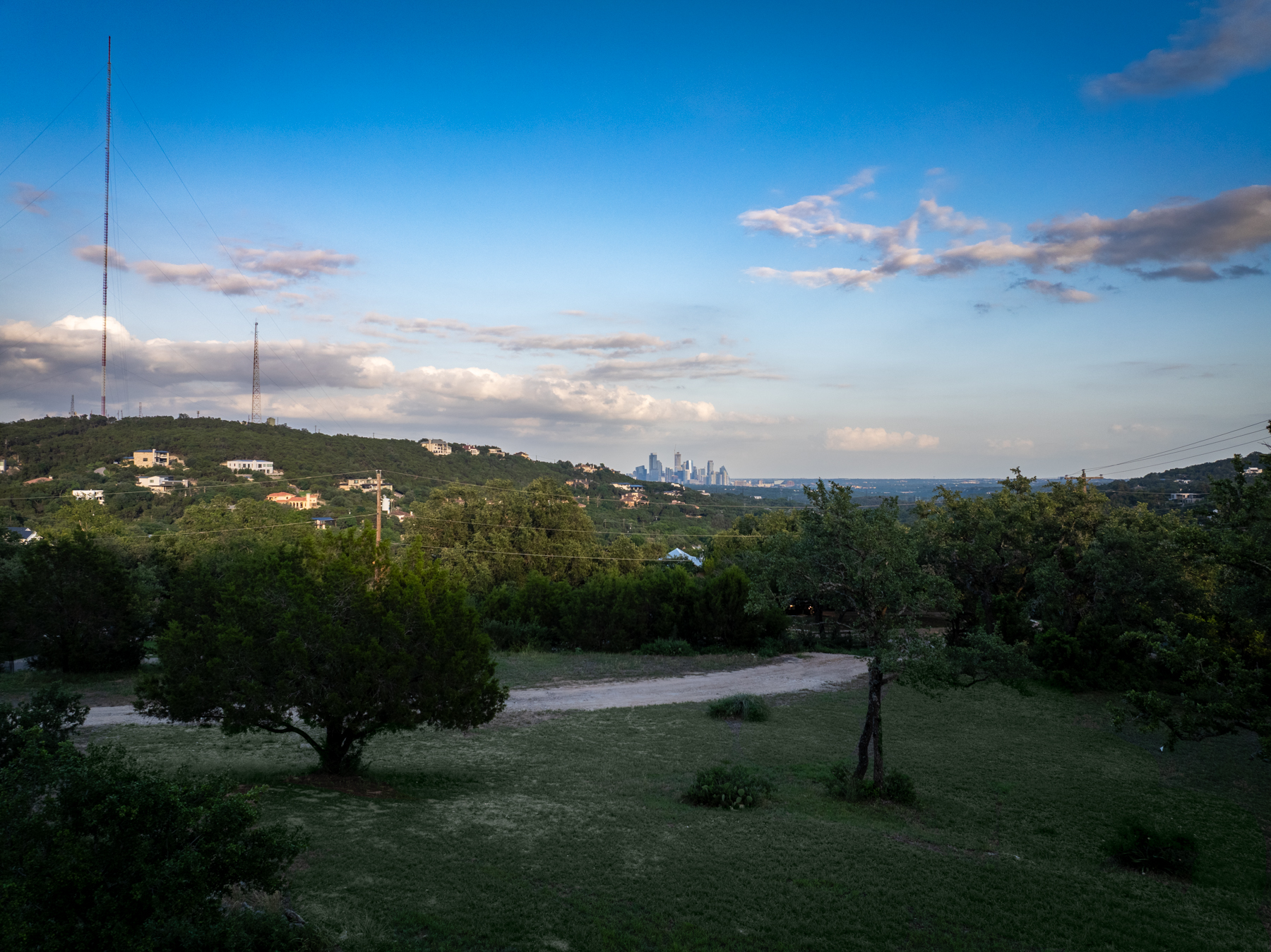 This image showcases a scenic, expansive backyard with a natural, gently sloping lawn and mature trees, highlighting the serene outdoor space of the property. In the distance, a clear view of a city skyline provides a dramatic contrast against the rolling, wooded hillside. The setting offers a peaceful atmosphere with significant potential for outdoor living and appreciation of scenic vistas.