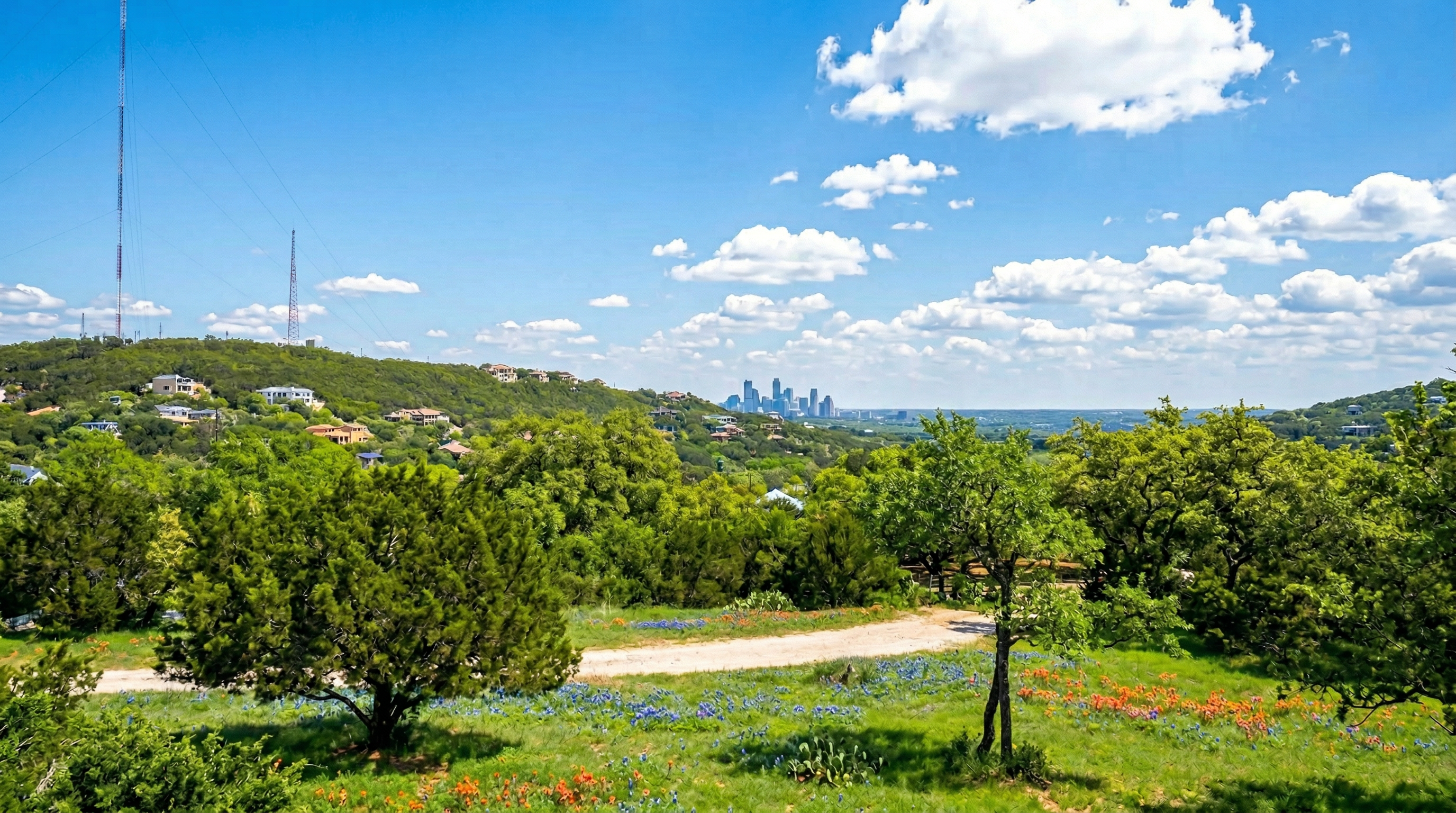 This scenic landscape photograph captures a hillside view overlooking a valley with the Austin skyline visible in the distance. The foreground features a vibrant field filled with wildflowers, including bluebonnets and Indian paintbrush, surrounding a dirt path. The hilly terrain is covered with lush trees and dotted with residential homes, emphasizing the picturesque and hilly nature of the location.