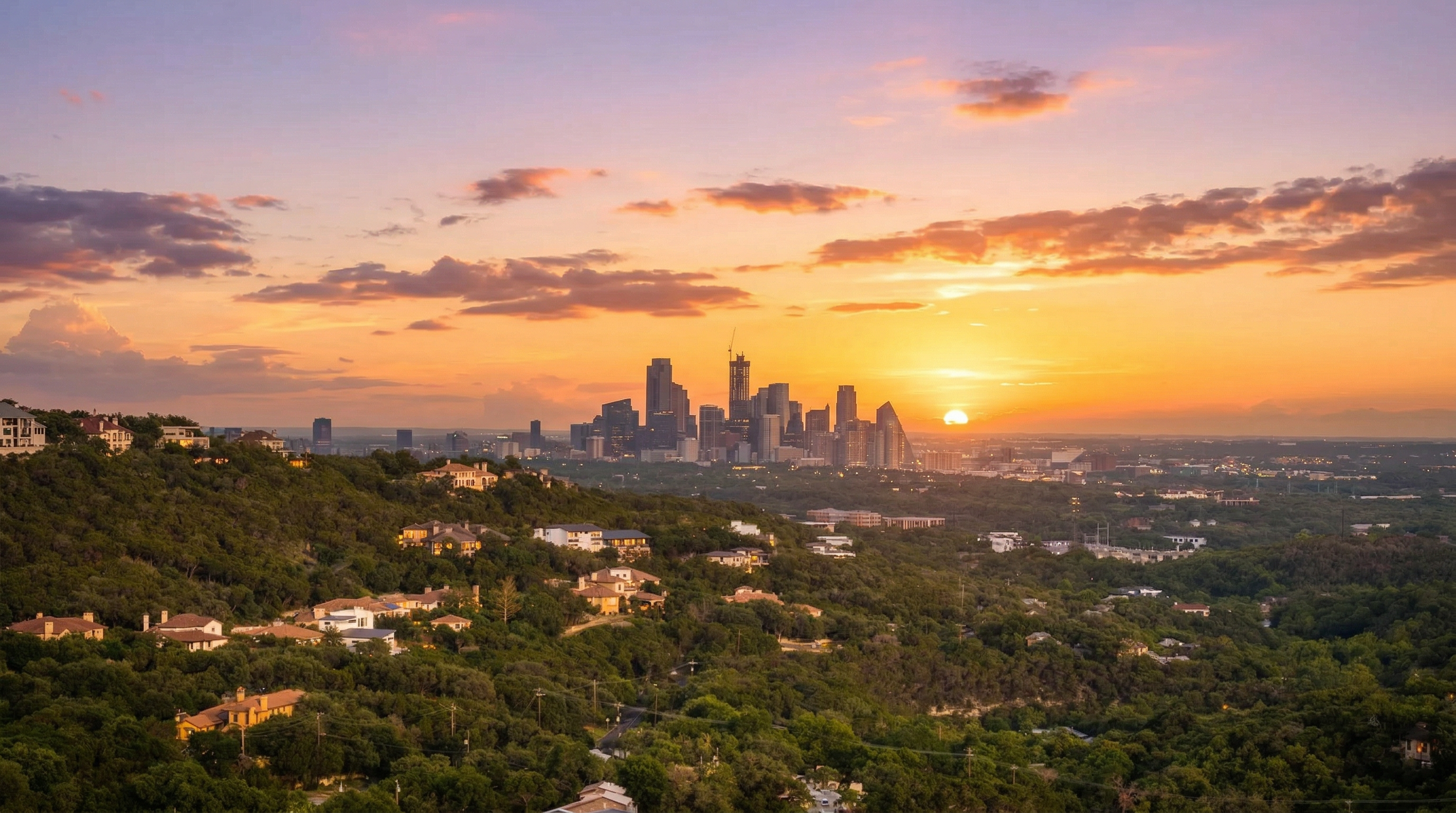 This panoramic aerial view captures the city skyline at sunset, set against a backdrop of rolling, tree-covered hills. The hillside features several well-appointed luxury residential homes nestled within the dense greenery, offering residents scenic views and a tranquil suburban setting. The image highlights the desirable blending of nature and urban proximity, characteristic of a premium real estate location.