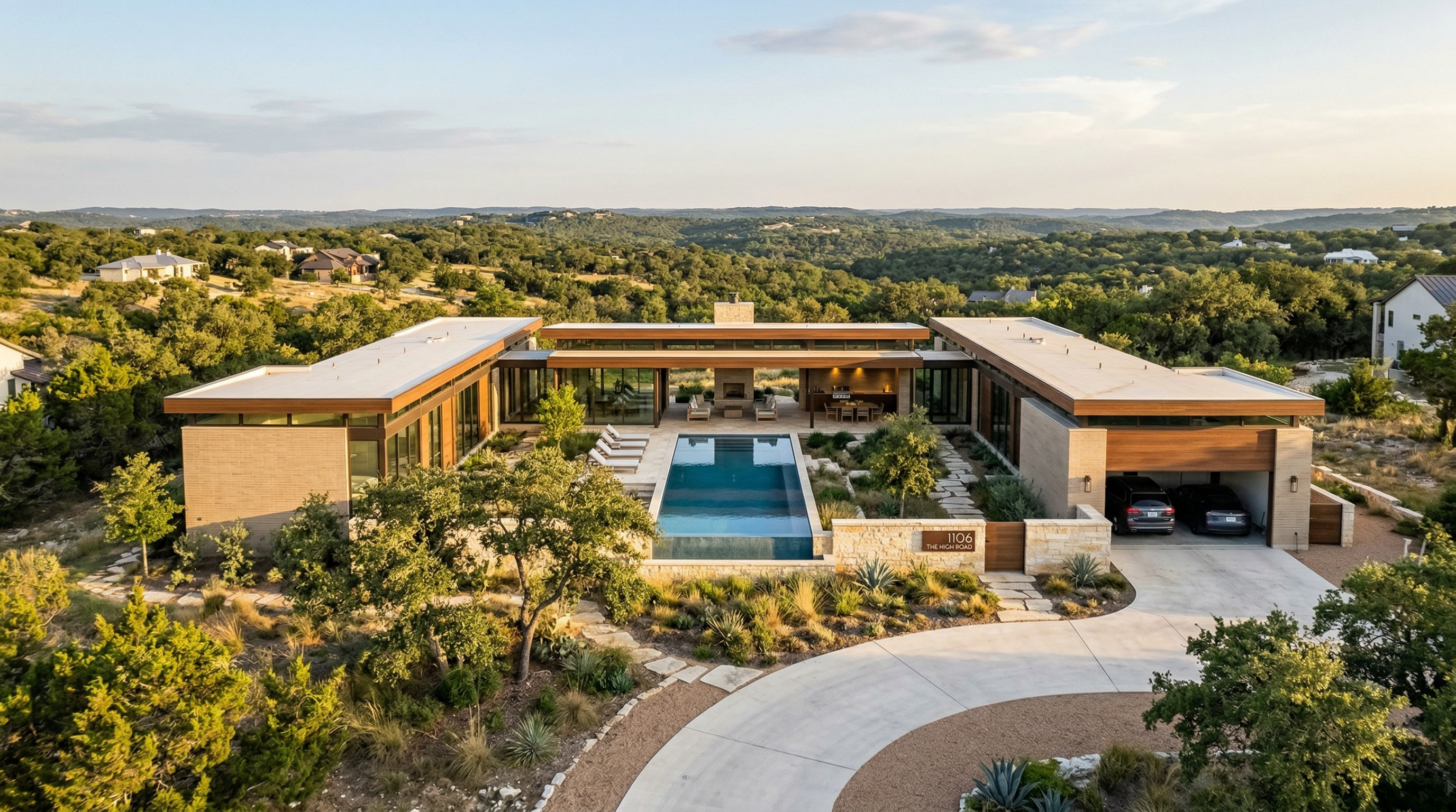 This stunning contemporary residence is captured in an expansive aerial view, showcasing its clean architectural lines and unique U-shaped design. The property features a centerpiece swimming pool, seamless indoor-outdoor living spaces, and a beautifully landscaped courtyard, all set against a tranquil, natural landscape. With modern garage bays, high-end stone finishes, and meticulous attention to site integration, this home offers a sophisticated, resort-like luxury aesthetic.