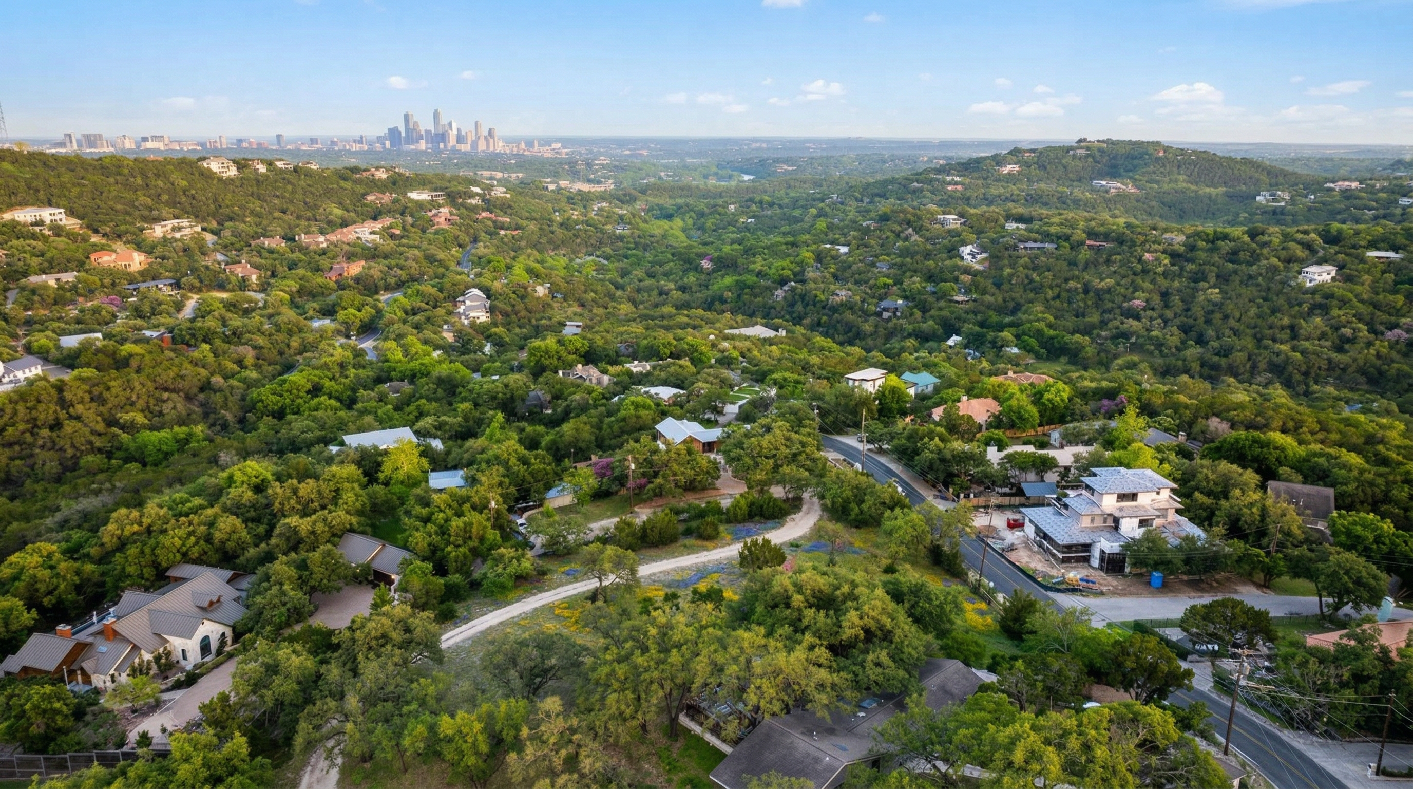 This high-altitude aerial view captures a sprawling, densely wooded residential neighborhood nestled on hills surrounding an urban city skyline. The landscape highlights several large, multi-story single-family homes integrated into the lush green terrain, with a winding road network and a mix of completed properties and active construction. It offers a prime example of scenic, secluded hillside living with convenient proximity to a major metropolitan area.