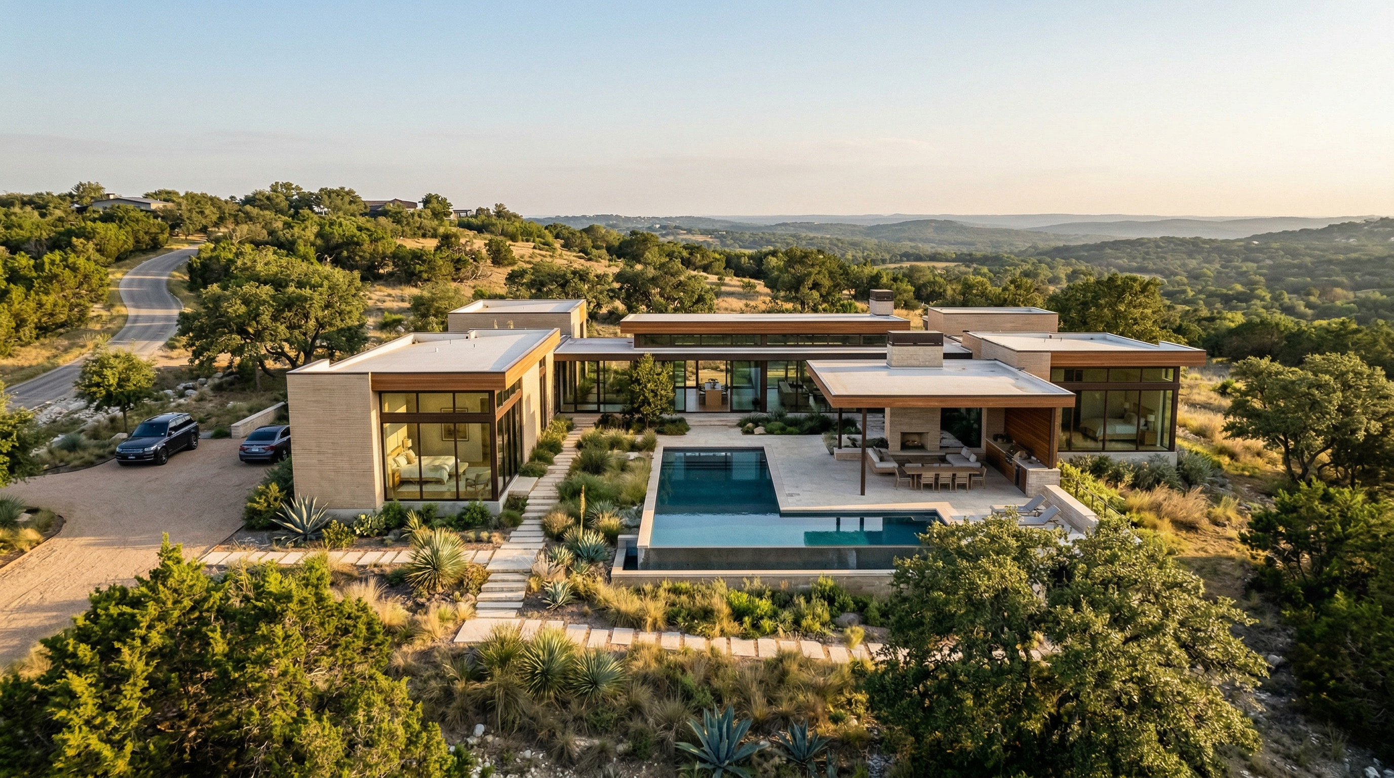 This stunning contemporary estate features a sprawling, low-profile architecture integrated into a rugged hillside landscape. The home centers around an L-shaped infinity pool and a generous outdoor patio with a fireplace, blending indoor and outdoor living seamlessly through large glass walls. Surrounded by native vegetation and rolling terrain, the property offers a sophisticated, minimalist aesthetic that emphasizes privacy and connection to the natural environment.