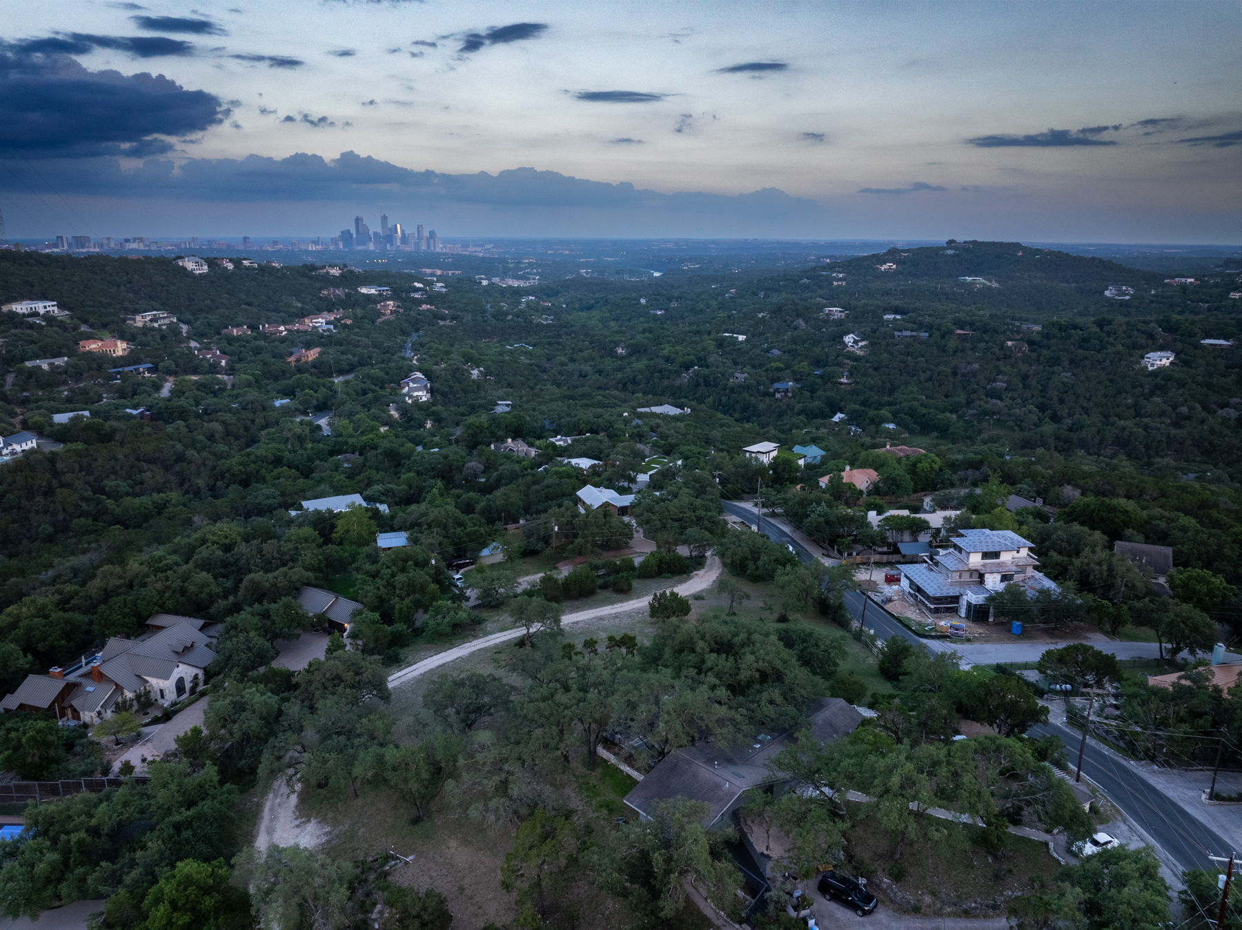 This panoramic aerial view captures a quiet hillside neighborhood nestled among dense, lush greenery. In the distance, the Austin, Texas skyline is visible against a moody, dusk-colored sky. The setting emphasizes privacy and natural beauty, highlighting the spacious, elevated lots typical of this scenic residential landscape.