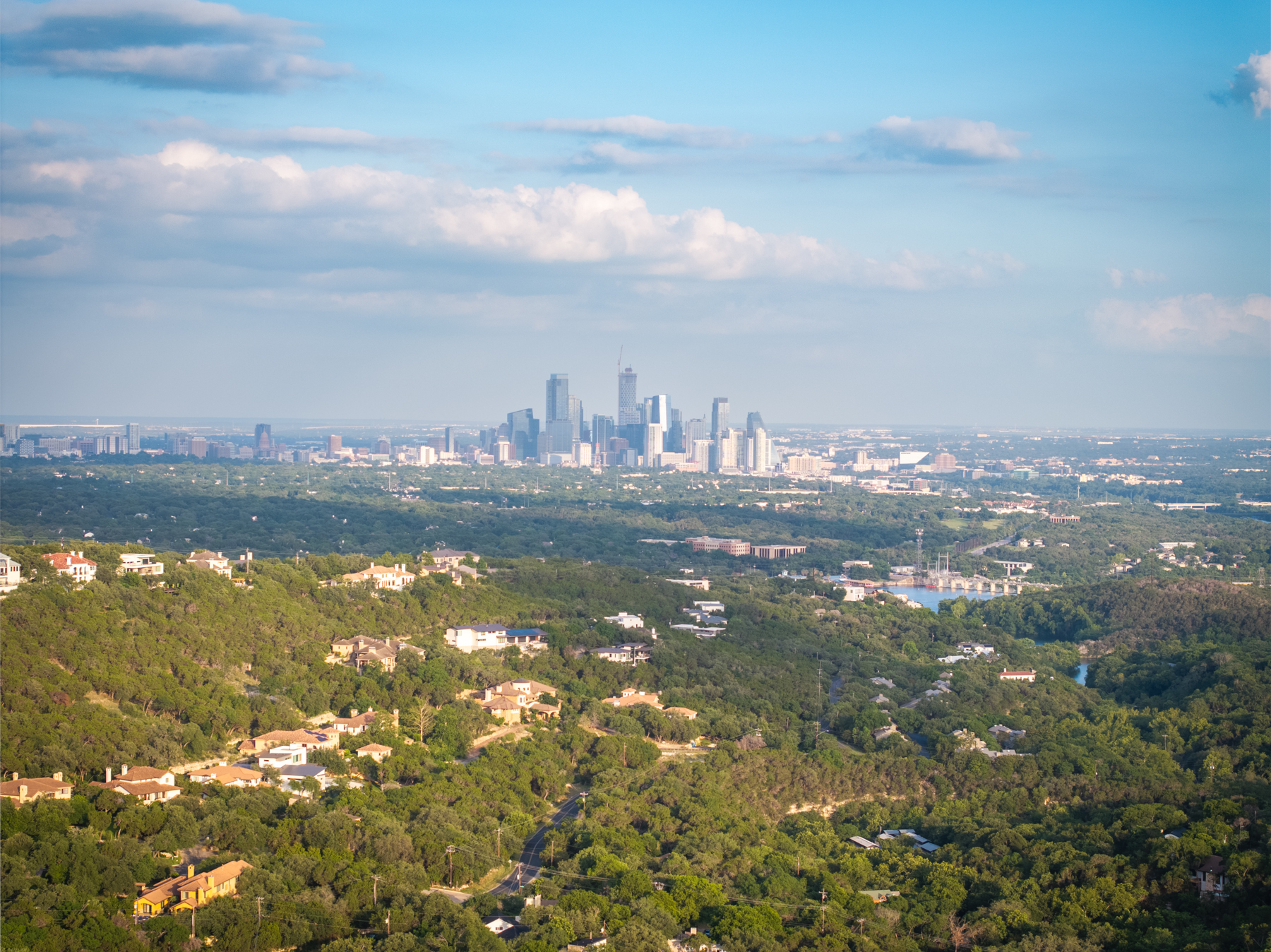 This stunning aerial view highlights a residential neighborhood nestled within a dense forest, offering a sense of seclusion and natural beauty. The foreground features custom-designed residential homes, while the background showcases a clear, panoramic skyline of a major city. This elevated vantage point provides high curb appeal by connecting luxury living with expansive urban convenience.