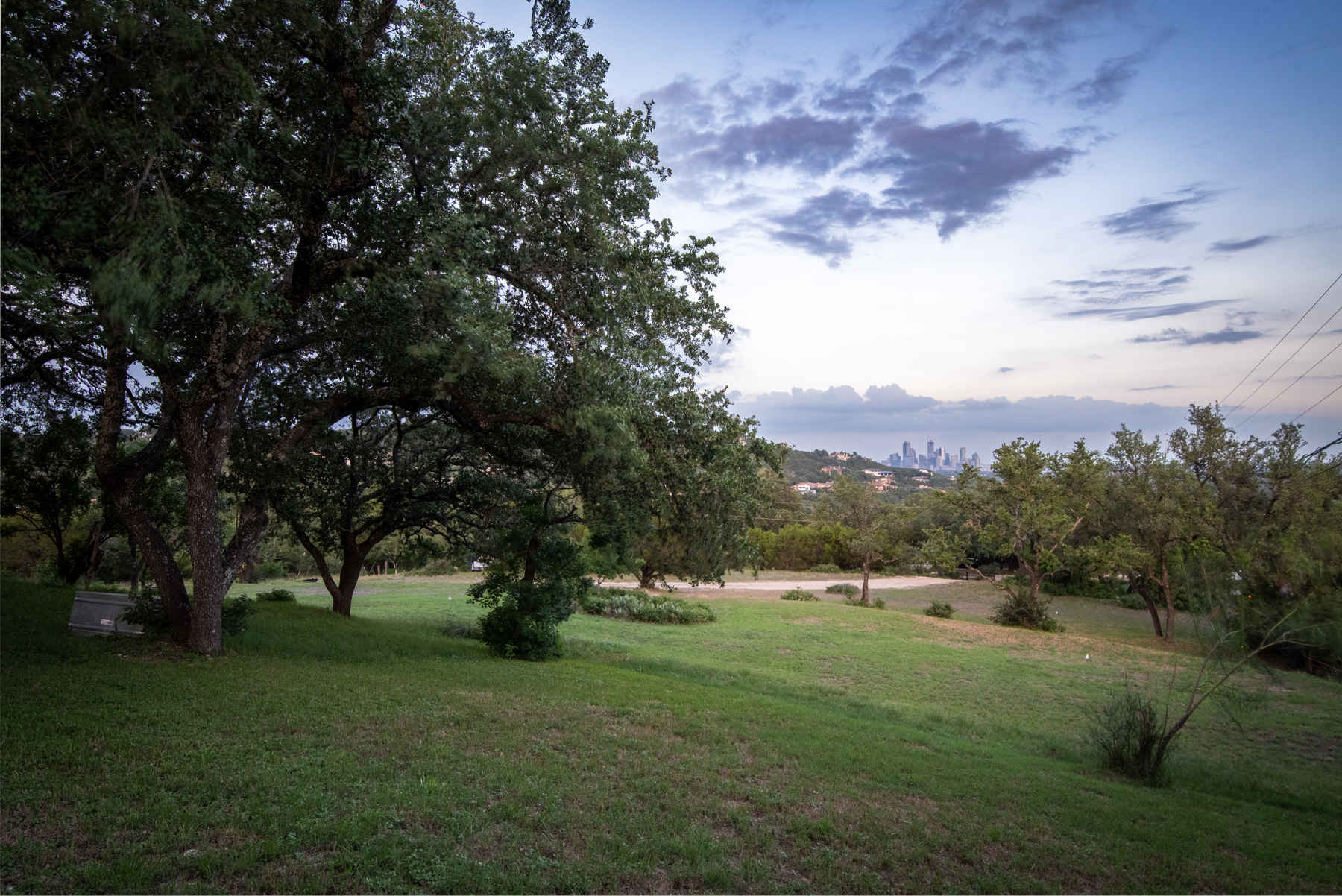 This expansive, grassy yard offers a peaceful natural setting with mature oak trees providing ample shade. The property features a gentle slope leading toward a scenic view of a distant city skyline under a soft evening sky. It is an ideal space for outdoor relaxation or entertaining, highlighting the property's connection to its tranquil, semi-rural surroundings.