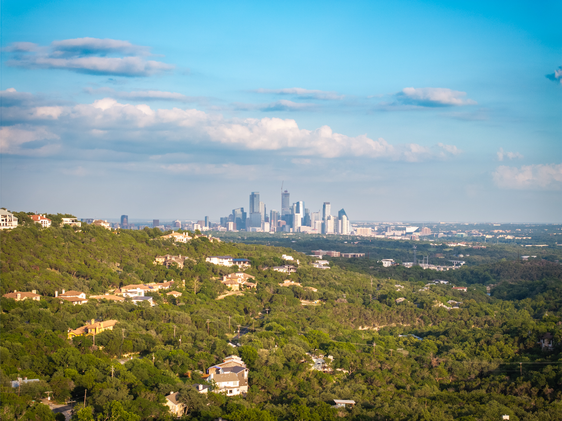 This panoramic aerial view captures a scenic, elevated perspective of a luxury residential neighborhood nestled within a lush, green landscape, leading the eye toward a prominent city skyline in the distance. The hillside terrain features winding roads and custom homes with architectural prominence, offering a serene and exclusive living environment. The clear blue sky and abundant mature trees enhance the appeal, showcasing an ideal setting that combines natural beauty with proximity to urban amenities.