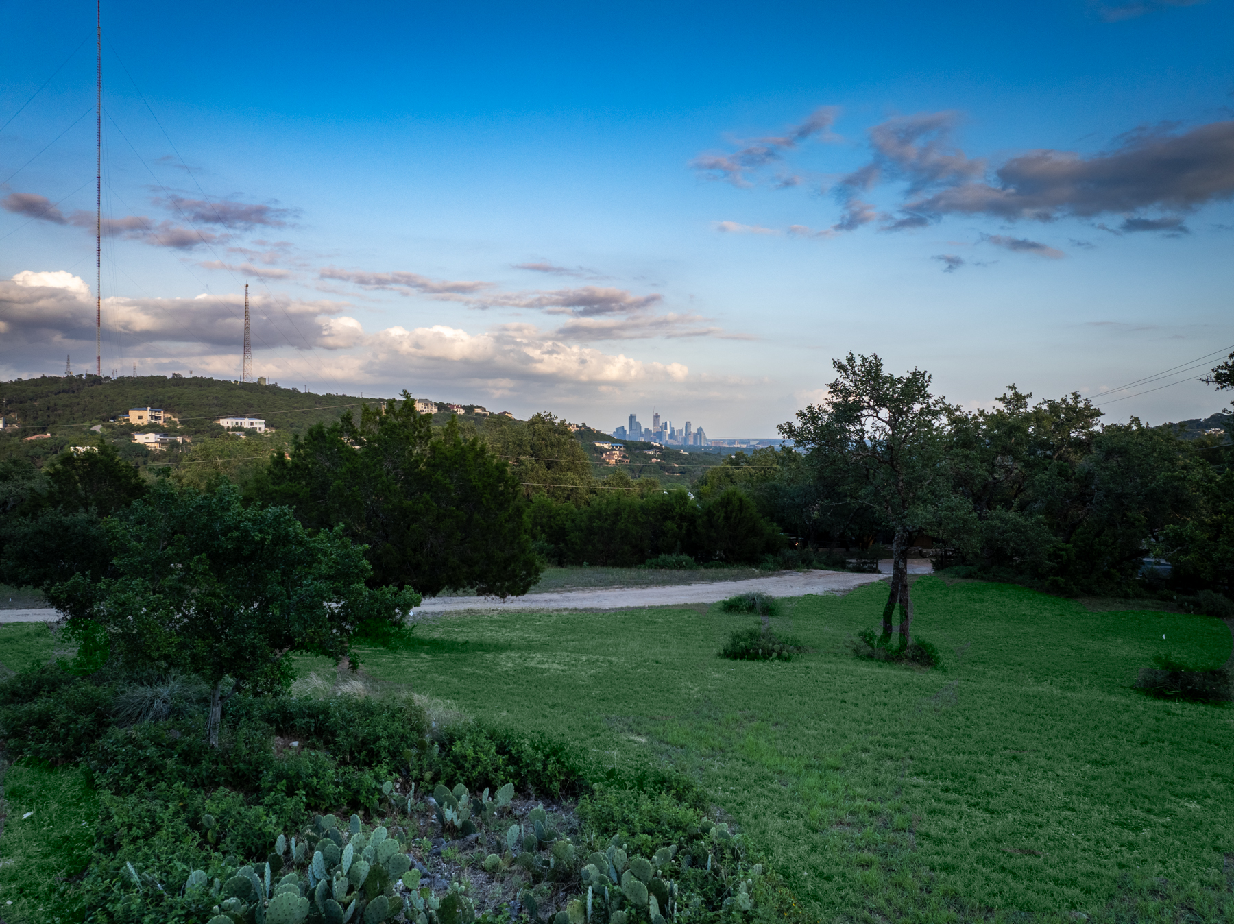 This panoramic view captures a lush, multi-tiered hillside landscape featuring native vegetation, including prickly pear cacti in the foreground and mature trees across the property. The expansive grassy yard leads the eye toward the rolling hills in the distance, which provide a stunning, unobstructed view of the distant urban skyline. It is an ideal outdoor space offering both natural serenity and a desirable scenic backdrop for potential homeowners.