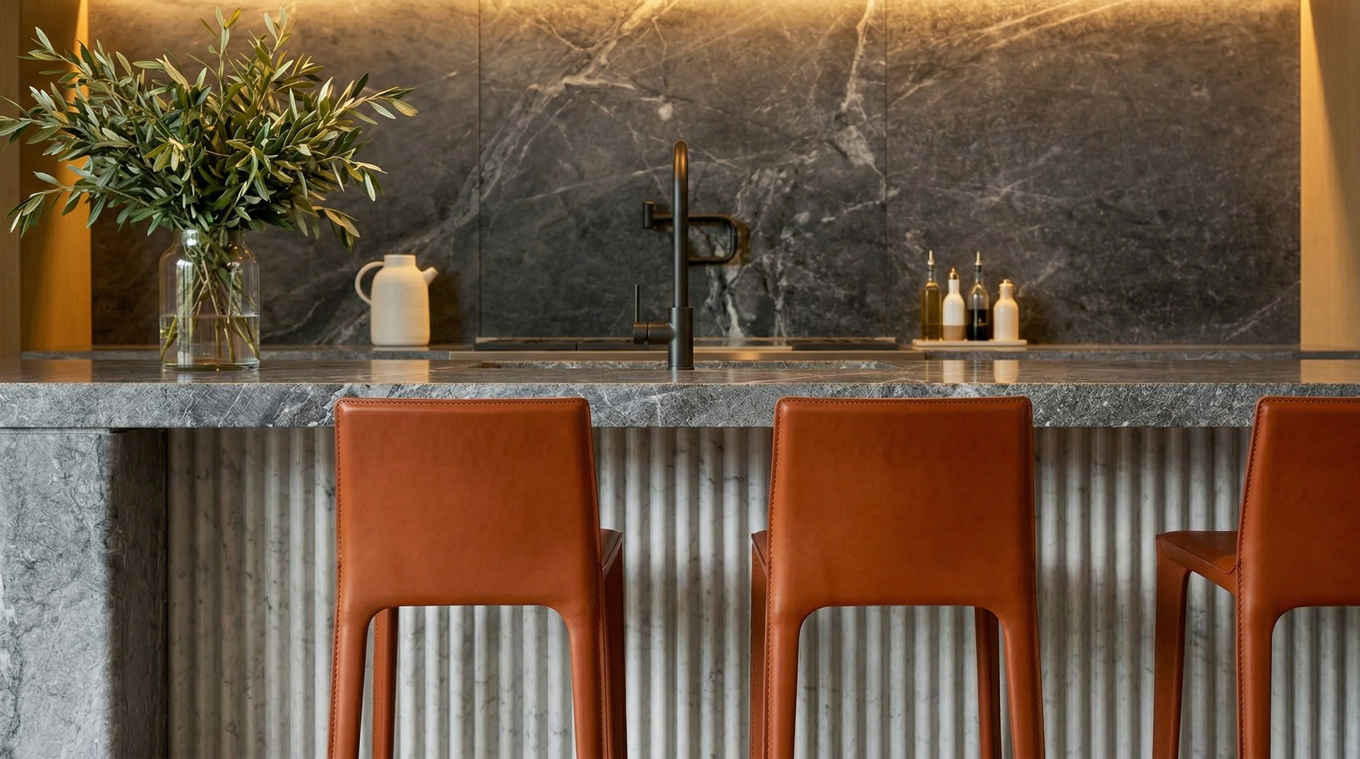 This sophisticated kitchen features a grand stone island with integrated sink and matte black faucet, paired with high-end, rust-colored leather bar stools. The backdrop is defined by dark, dramatic marble slab backsplashes and vertical ridged marble detailing, creating a luxurious and modern aesthetic. The warm, concealed under-cabinet lighting adds a welcoming ambiance to this high-end culinary space.