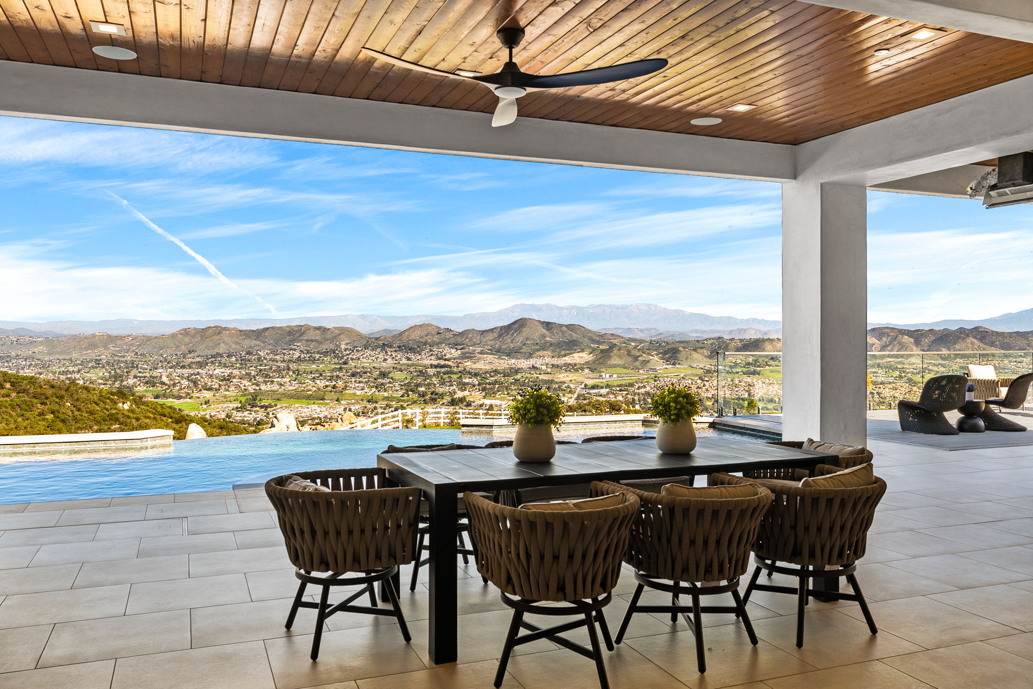 This stunning covered patio area showcases an infinity-edge pool that seamlessly blends with the panoramic valley and mountain views. The space is elegantly furnished with a modern dining set featuring woven chairs and features a wooden ceiling with a large contemporary ceiling fan. The overall atmosphere is luxurious and ideal for outdoor entertaining with a picturesque backdrop.