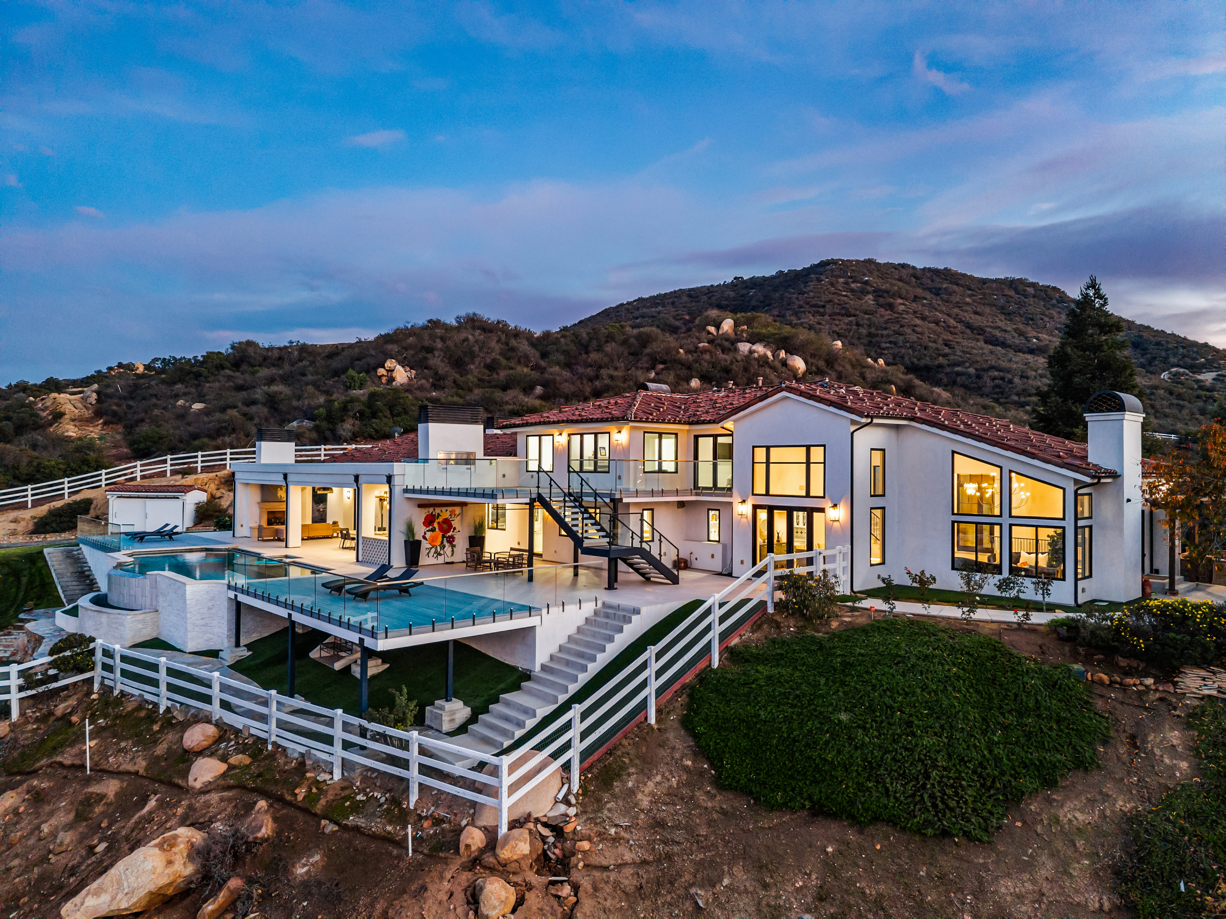 This stunning luxury estate is captured from an elevated perspective, showcasing a modern architectural design with a spacious multi-level deck and an infinity-edge pool. The property features a crisp white facade, large picture windows, and a beautifully integrated hillside landscape with a white fence. Its elevated position and seamless indoor-outdoor living spaces create an impressive and sophisticated residential aesthetic.
