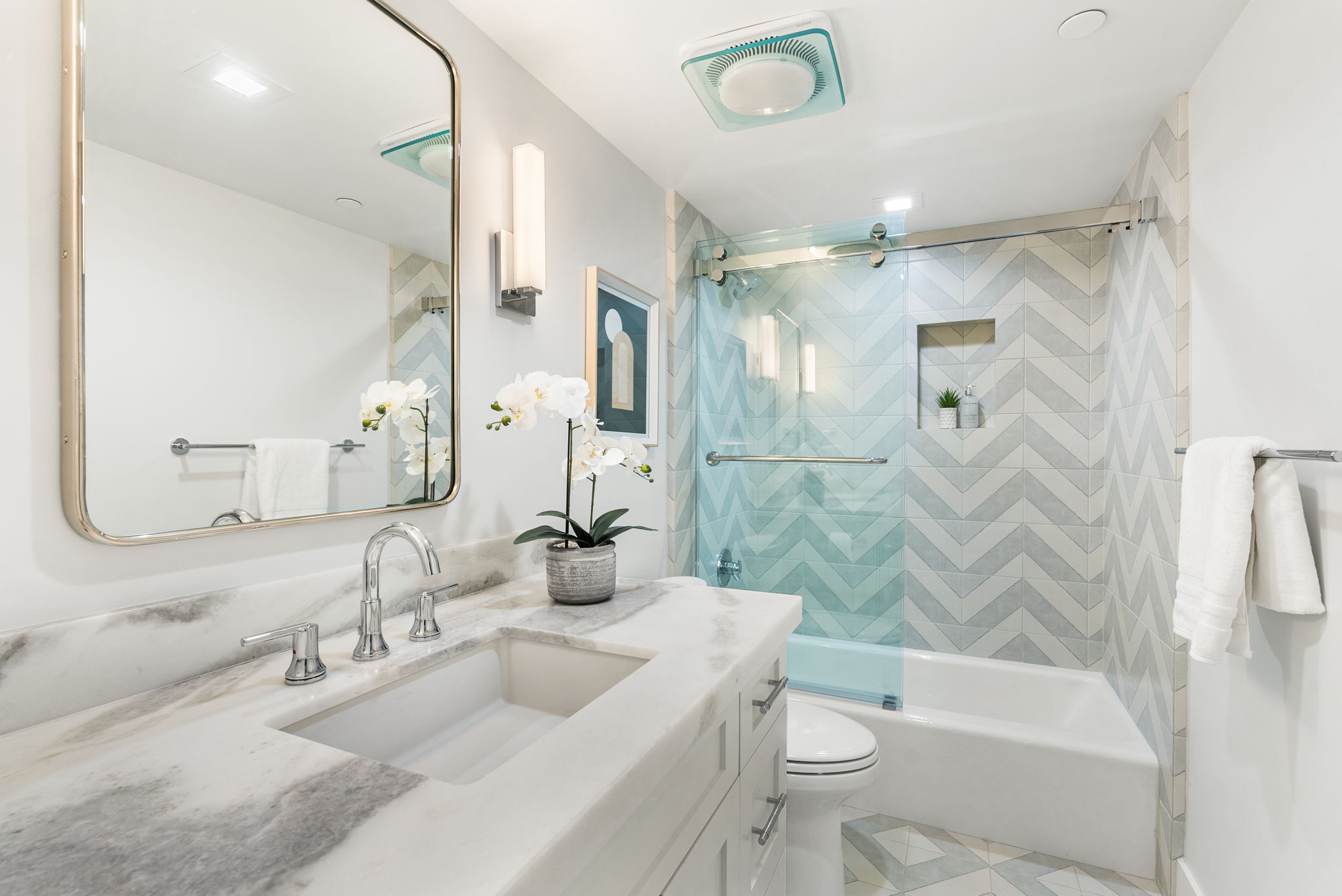 This modern, bright bathroom features a clean white vanity with a marble countertop and a striking chevron-patterned tiled tub surround in soft blue and gray tones. A sleek glass sliding shower door and gold-framed wall mirror add sophisticated, contemporary accents to the space. The room is well-lit and thoughtfully staged, presenting a polished and inviting aesthetic suitable for a guest or secondary bath.