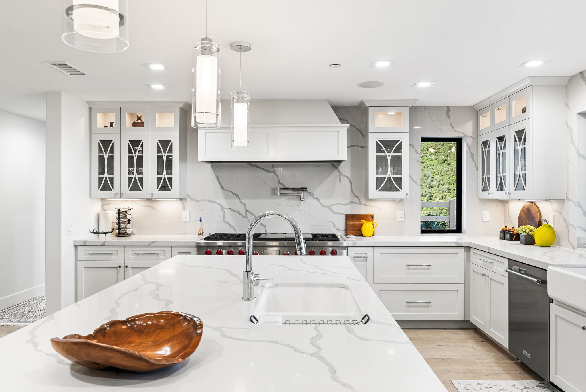 This modern kitchen features elegant white cabinetry paired with striking marble-patterned quartz countertops and a full-height matching backsplash. The spacious center island serves as a focal point, complemented by stylish pendant lighting and high-end stainless steel appliances, including a professional-grade range. With its bright, clean aesthetic and a window providing natural light, the space creates an inviting and sophisticated atmosphere perfect for cooking and entertaining.