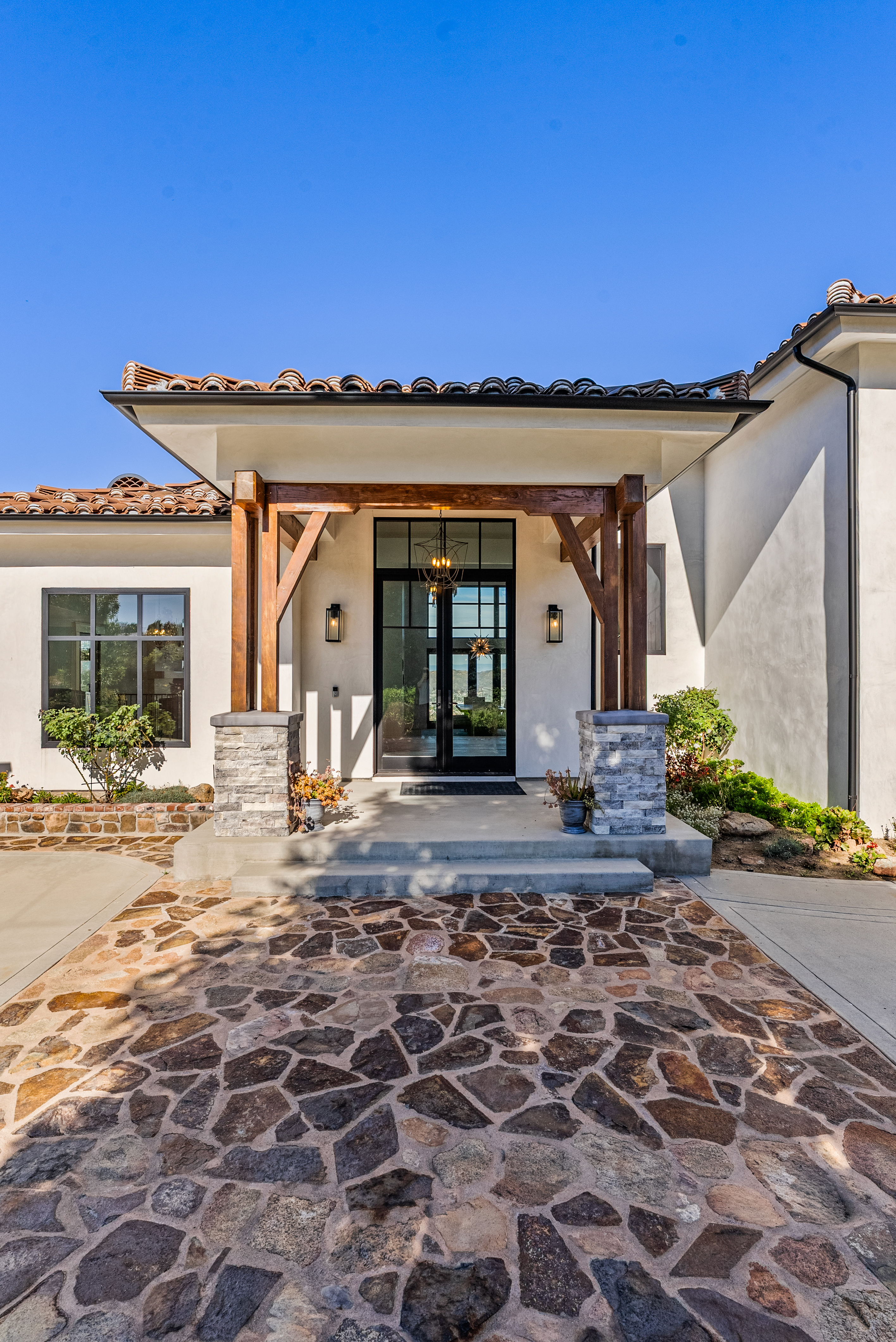 This elegant home features a welcoming entryway with a prominent covered porch, defined by sturdy wooden posts set on stacked stone bases. The facade is characterized by clean white stucco walls, decorative Spanish-style clay roof tiles, and a sophisticated black-framed glass double door. The front of the property is completed by a charming rustic stone-paved walkway that leads directly to the inviting entrance.